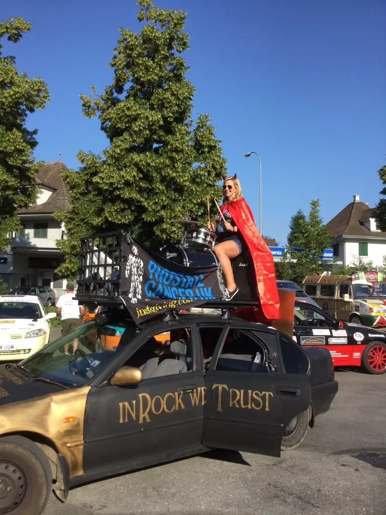 A person dressed as a rock star sitting on the roof of a decorated black car with gold accents, playing drums. The car has the phrase 'IN ROCK WE TRUST' painted on the side. There are other cars and people in the background, and the person is wearing