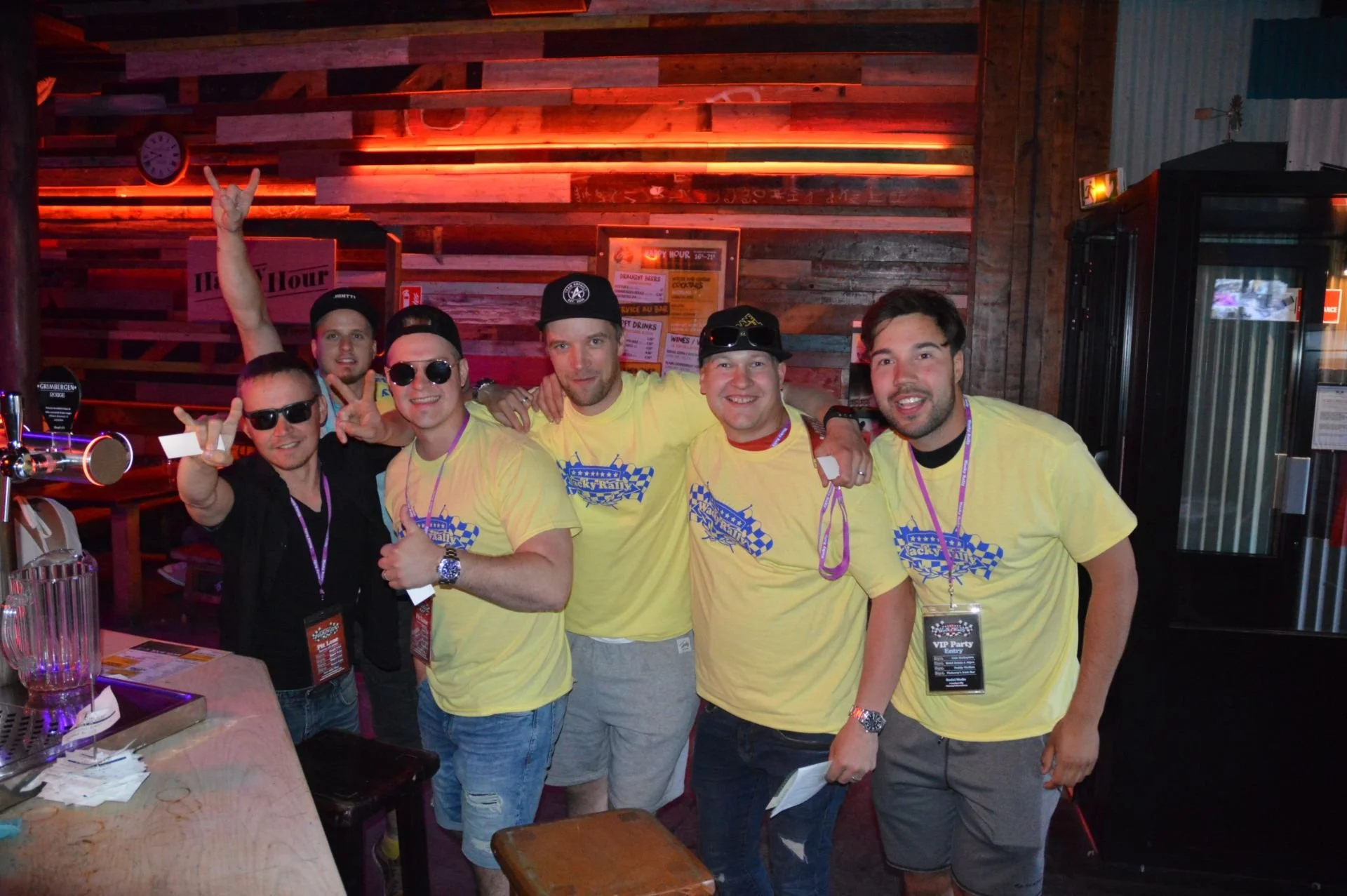 Group of six young men in matching yellow T-shirts with blue checkered flags, posing inside a bar or restaurant with wooden walls and neon lighting, some wearing sunglasses and lanyards.