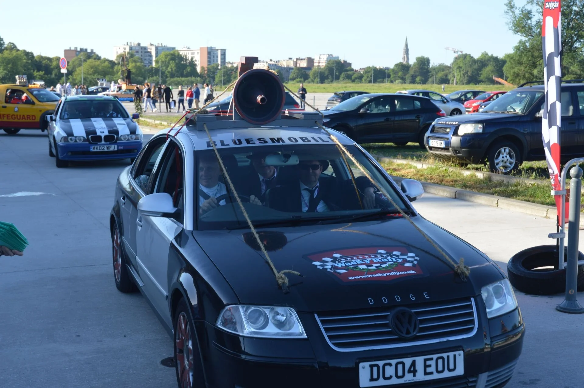 A silver Volkswagen car with a large megaphone on the roof and ropes attached, participating in a rally or parade called 'Bluesmobil.' Several other vehicles are visible in the background, including a police car, a yellow lifeguard vehicle, and a bla