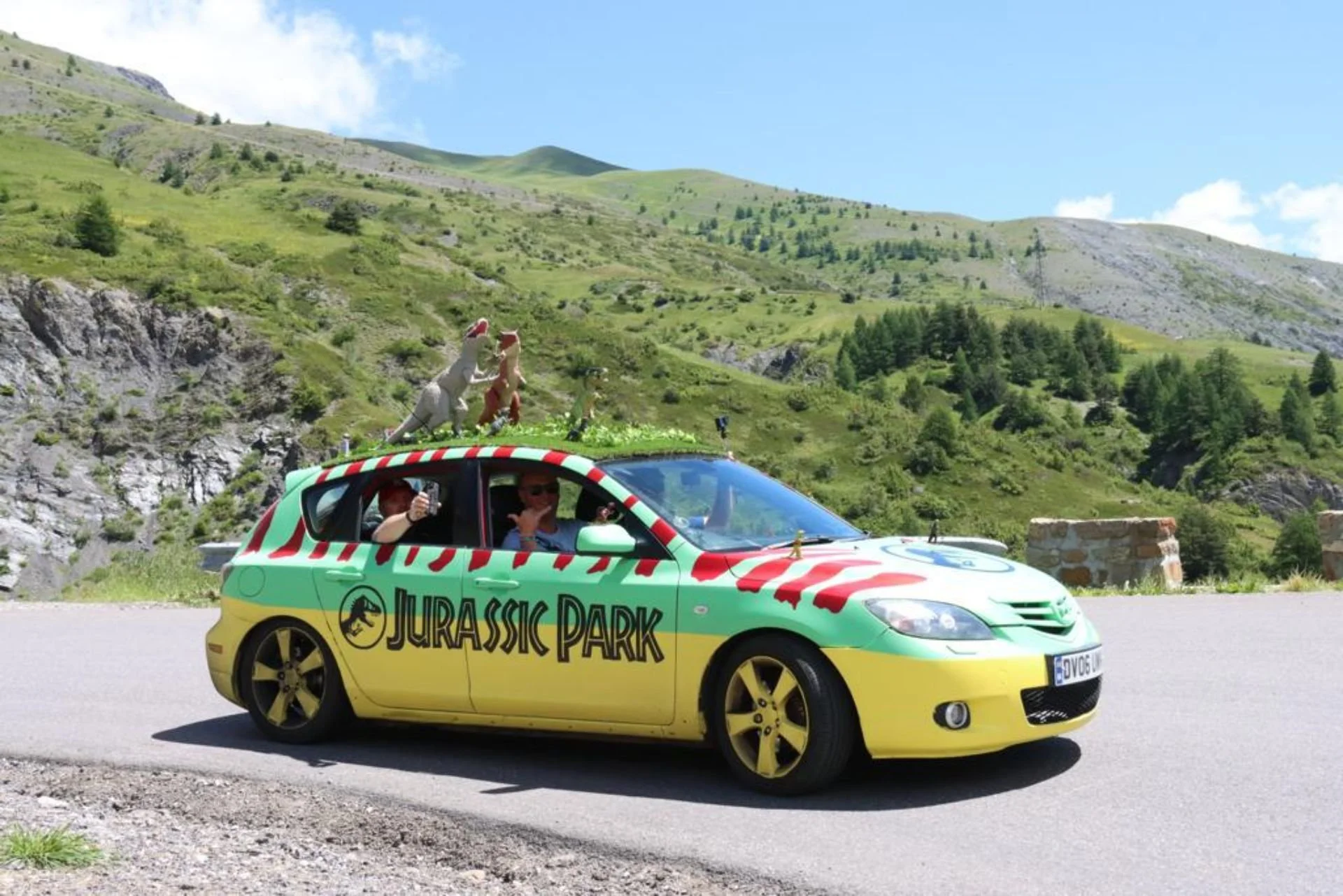 A decorated yellow and green car labeled 'Jurassic Park' with dinosaur figures on top, parked on a mountain road, with lush green hills and trees in the background.