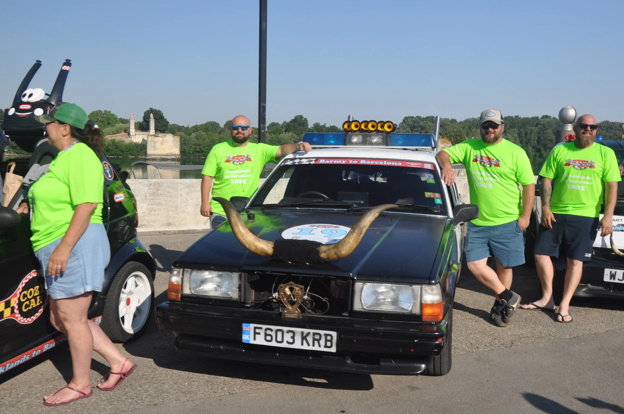 A group of people wearing bright green t-shirts standing next to a decorated black car with large horns attached to its front, parked near a waterfront with historic buildings in the background.