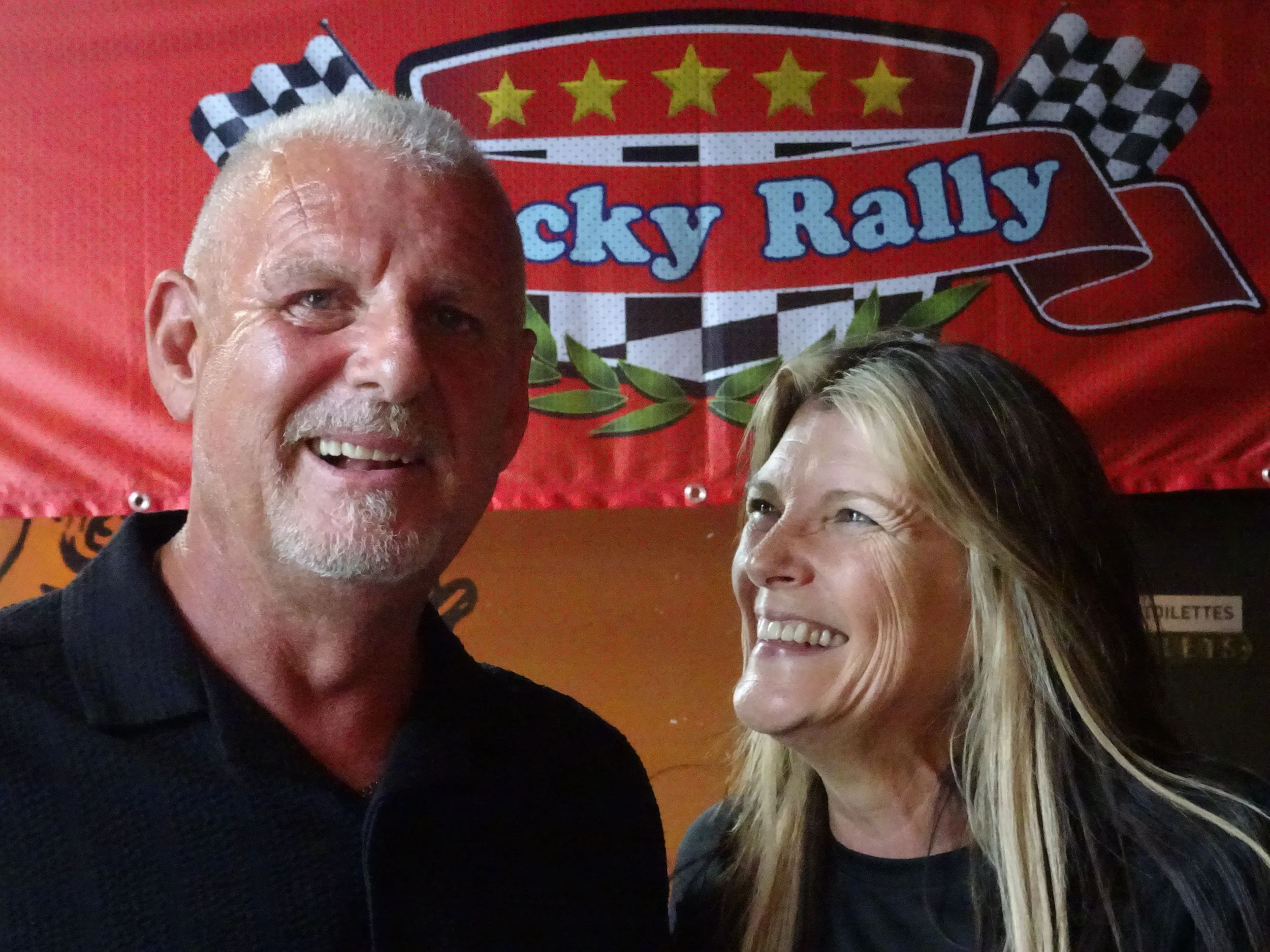 A smiling man with gray hair and a beard standing next to a smiling woman with long blonde hair. They are in front of a red backdrop with the words 'Lucky Rally' and checkered flags.