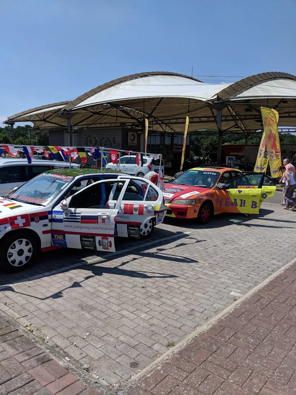 Colorful decorated cars parked outside a gas station with flags and banners, under a large canopy on a sunny day.