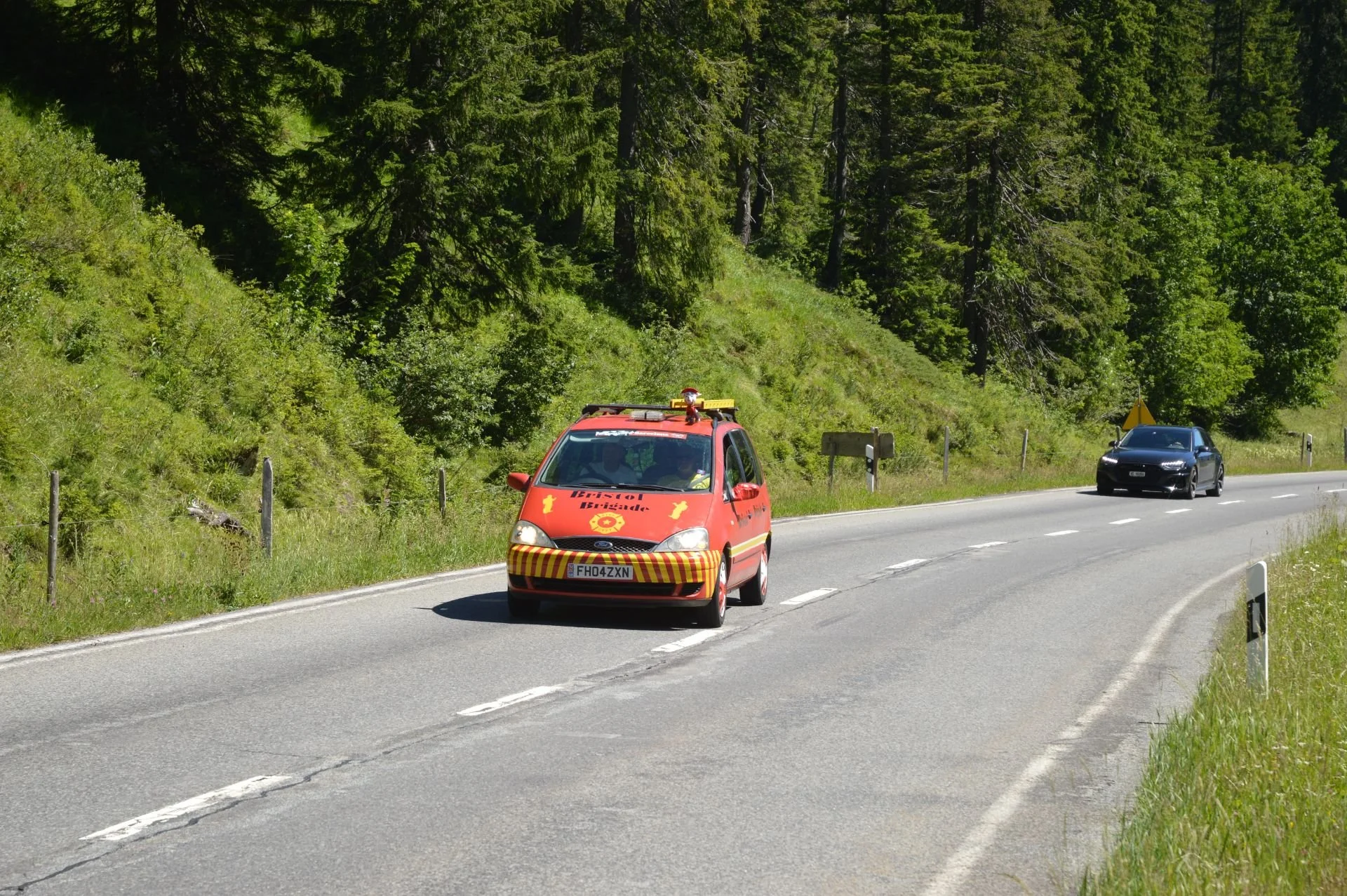 A red emergency vehicle with yellow markings and siren on top driving on a rural highway, with a black sports car following behind, surrounded by green trees and grass.