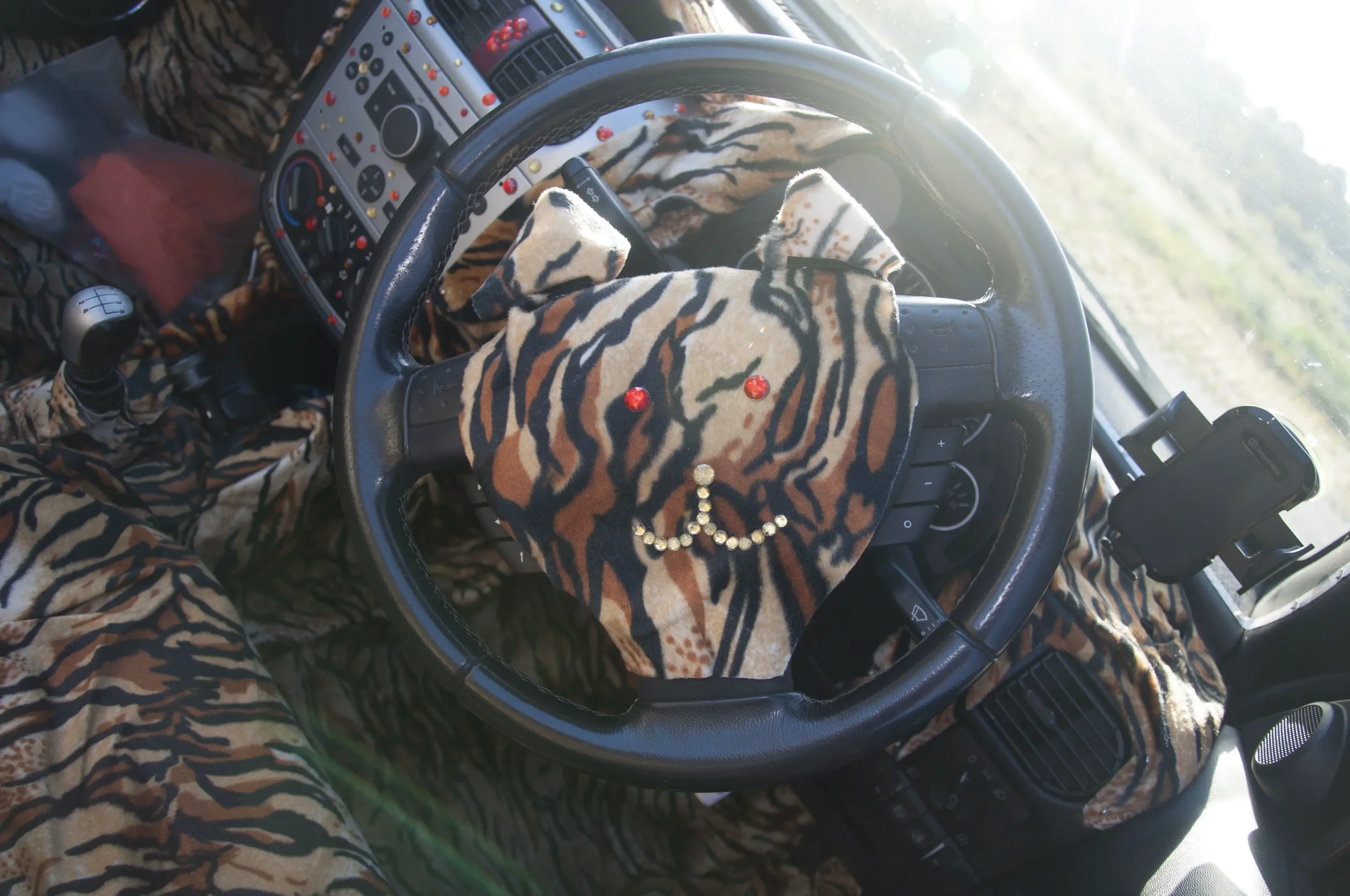 Interior of a vehicle with tiger print fabric covering the steering wheel, dashboard, and seats. The steering wheel has a plush tiger print cover with a peace sign made of rhinestones and small red decorations resembling cherries. A steering wheel ph