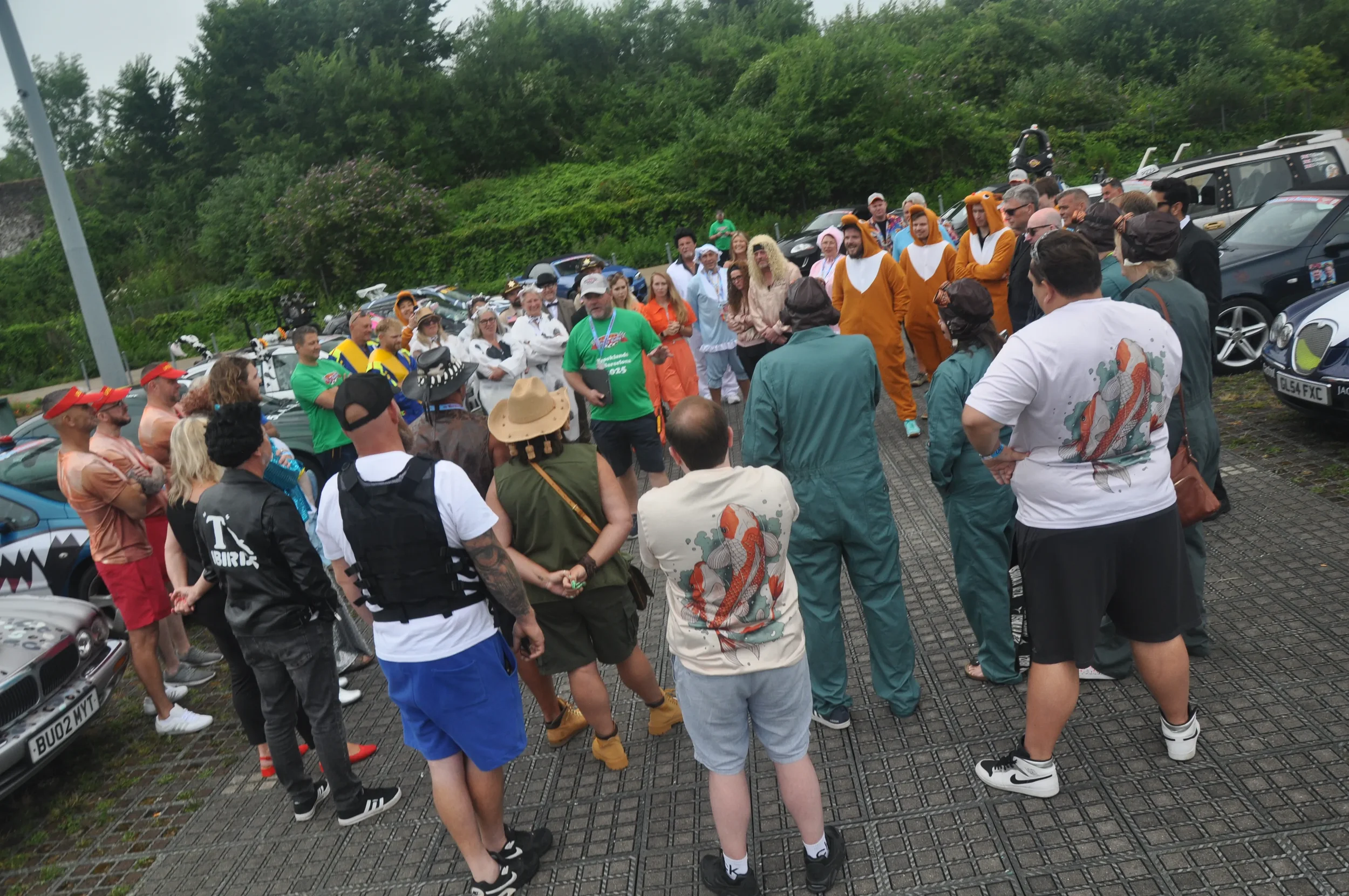 Group of people gathered outdoors in a parking lot, some dressed in colorful costumes including animal onesies and themed outfits, with a background of cars and lush green trees.