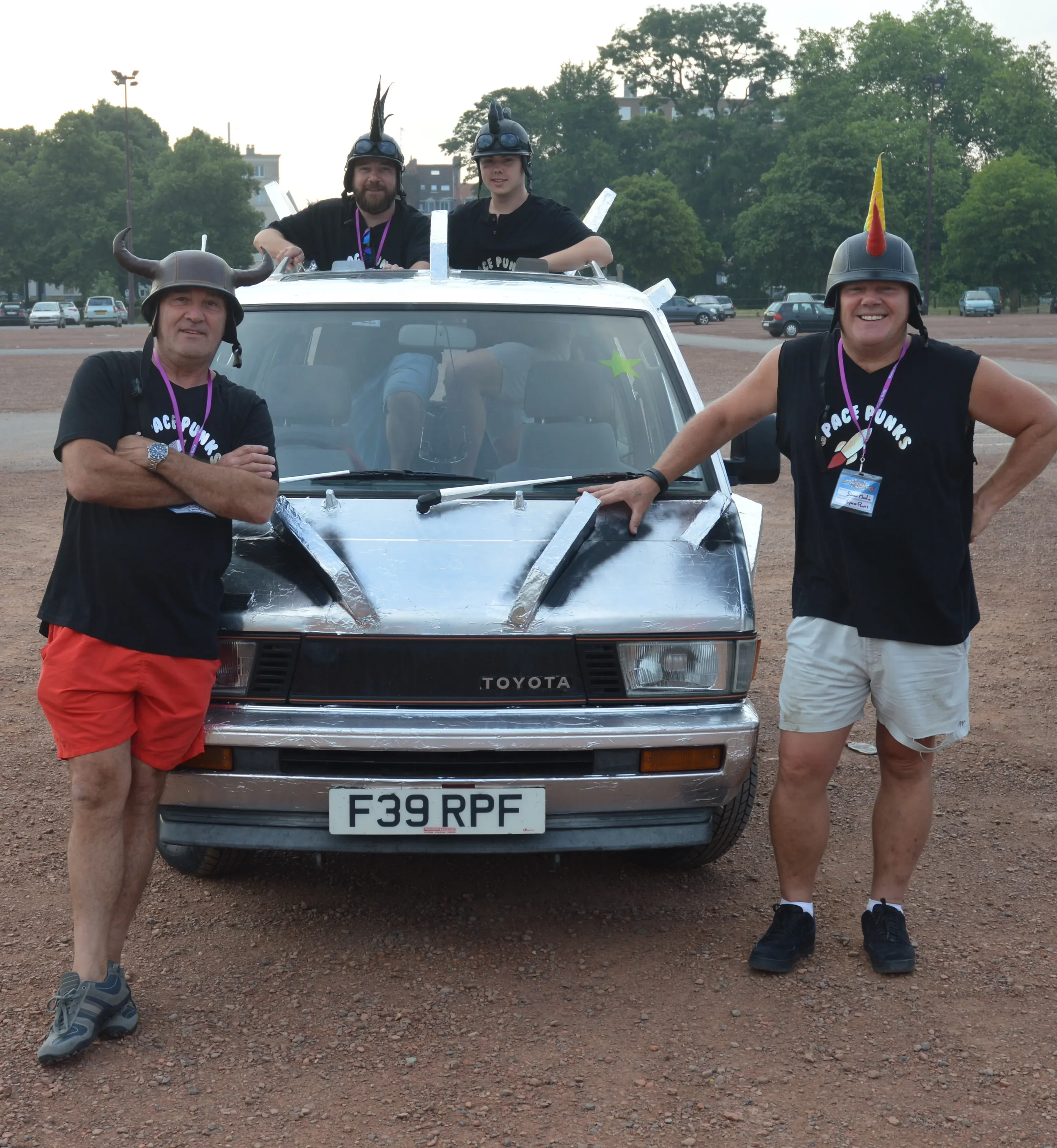 Four men wearing humorous helmets and black 'Space Punks' T-shirts, posing in front of and on a creatively decorated vintage Toyota car at an outdoor event in a parking lot.