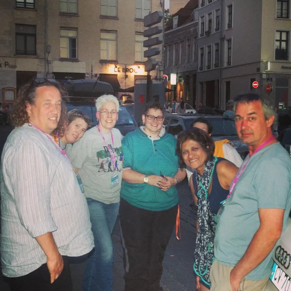 Group of seven people standing on a city sidewalk during the evening, smiling at the camera, with buildings and cars in the background.
