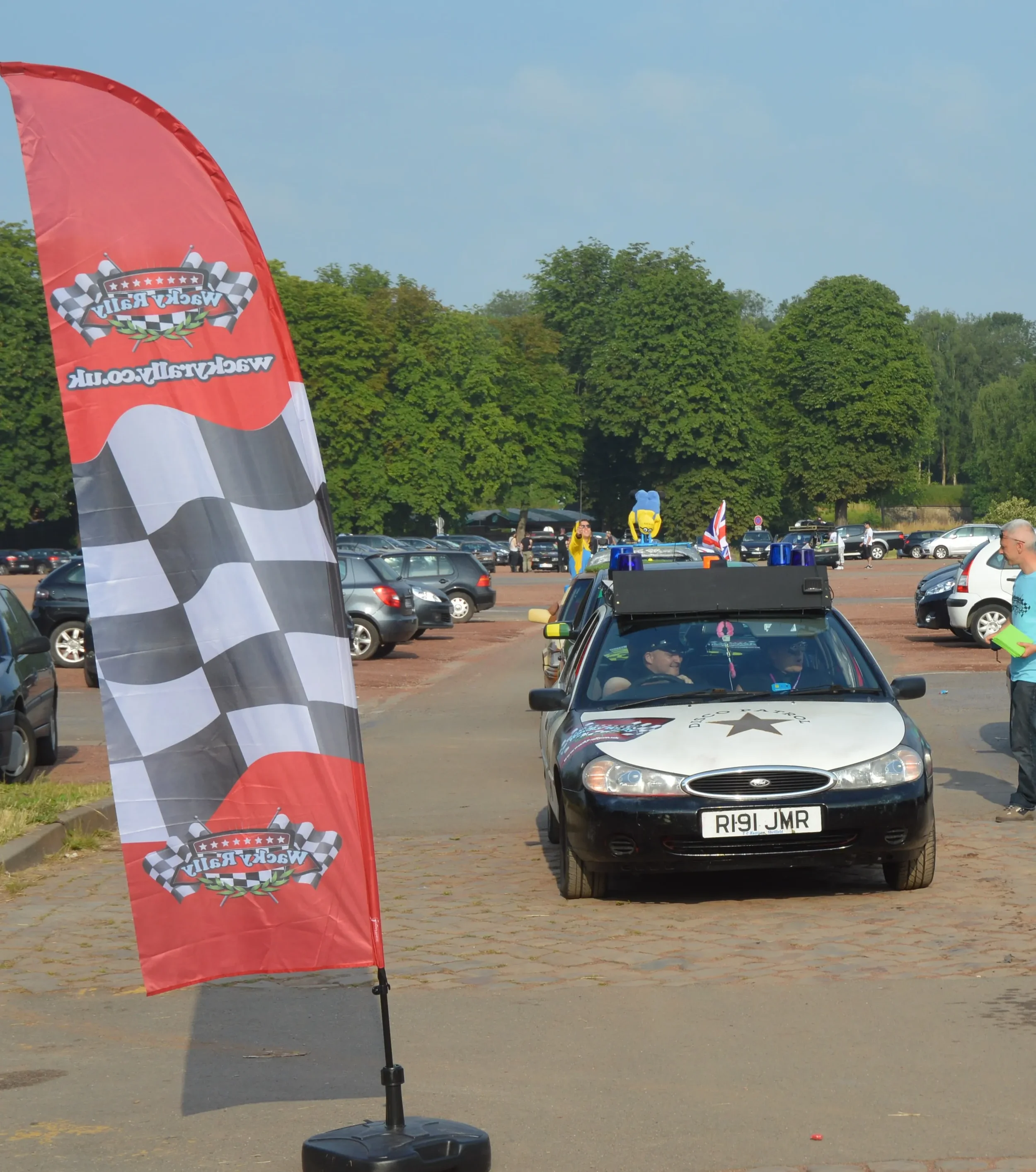 A service vehicle with a siren and Minion decorations on the roof parked in a parking lot during a racing event. There is a red and black checkered flag flag nearby and several other cars in the background.