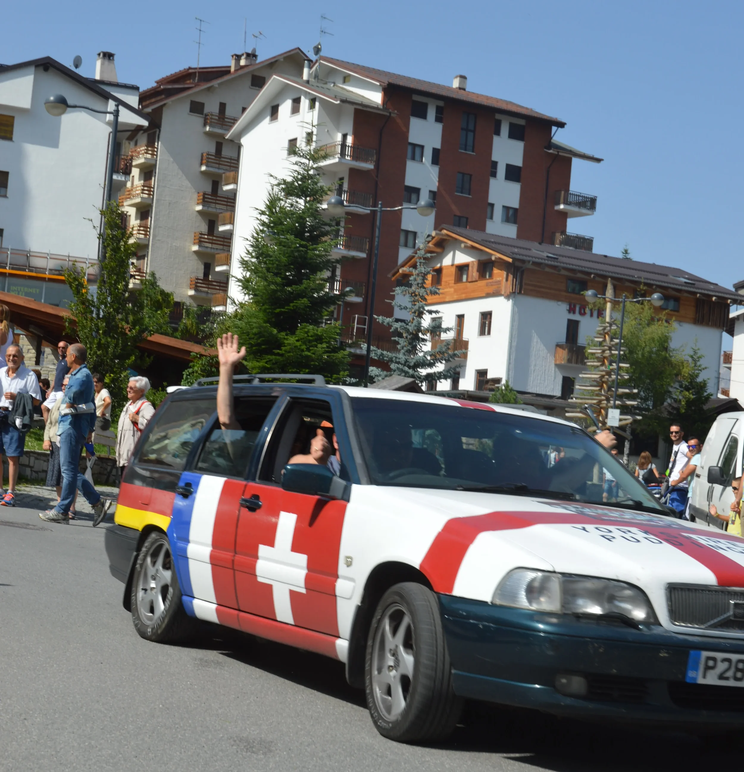 A car with Swiss cross and rainbow stripes on its side, driving in a parade, with people walking and waving on the sidewalk.