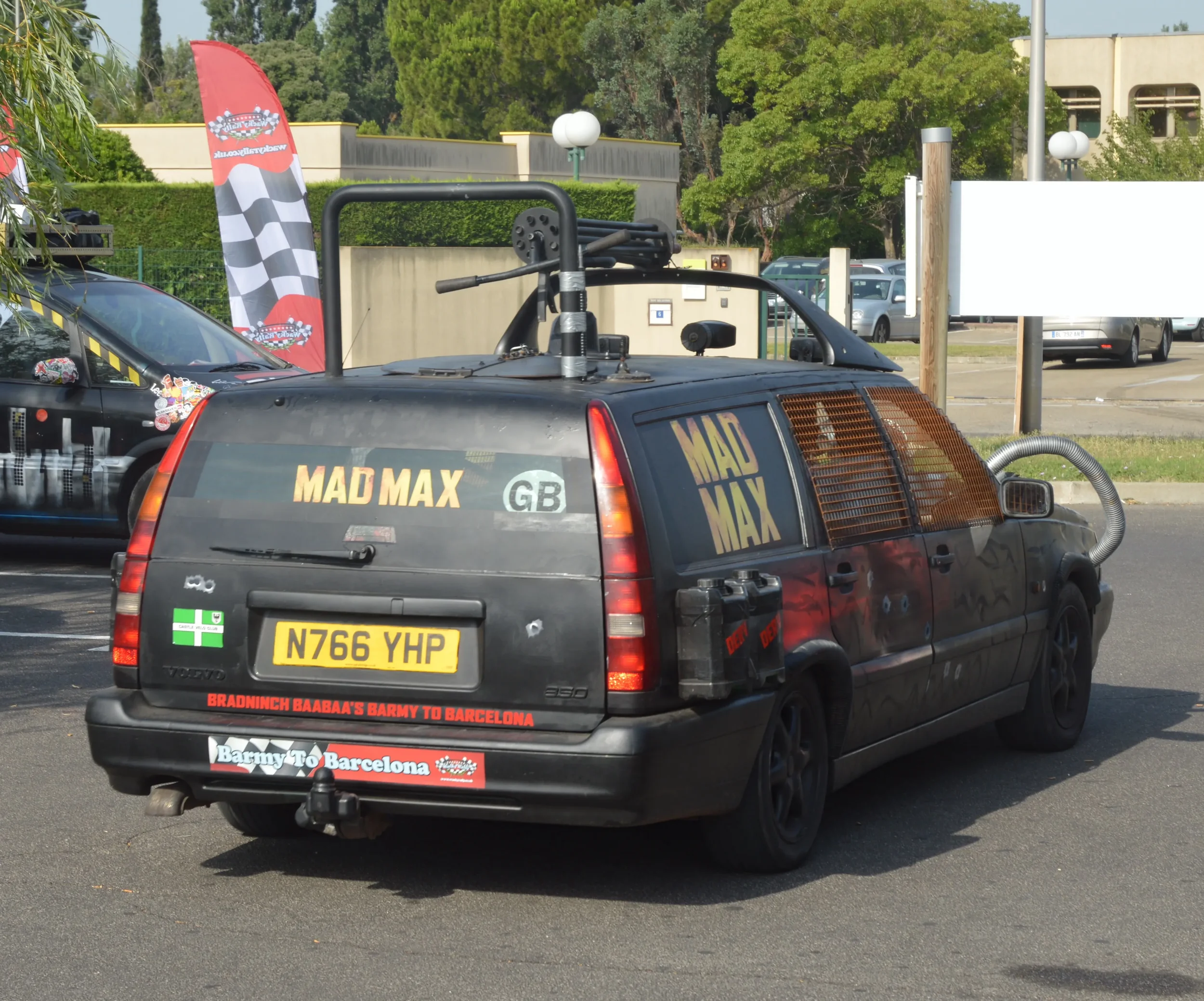 A black fictional vehicle from the Mad Max movies, parked in a lot. It has various stickers and modifications, including a yellow license plate (N766 YHP), large pipes on the roof, and a large exhaust pipe. There are additional stickers referencing B