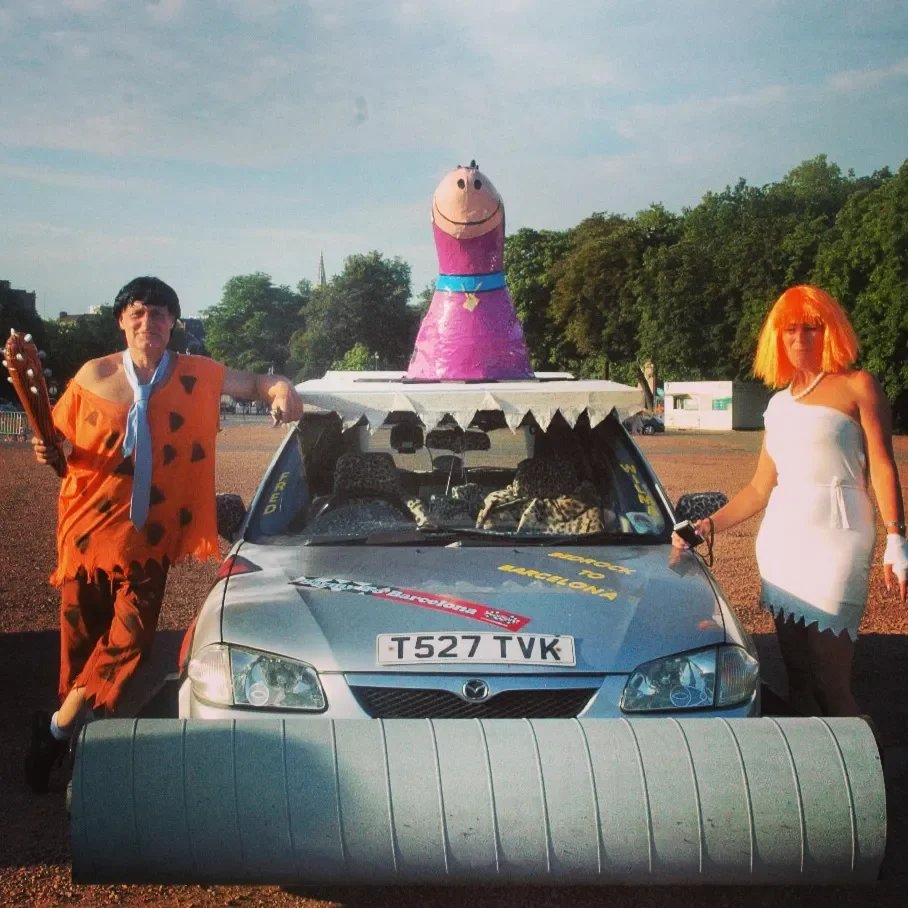 A person dressed as Fred Flintstone stands next to a decorated car with a large inflatable Pink Panther head on top, and another person dressed as Wilma Flintstone standing nearby, at an outdoor event.