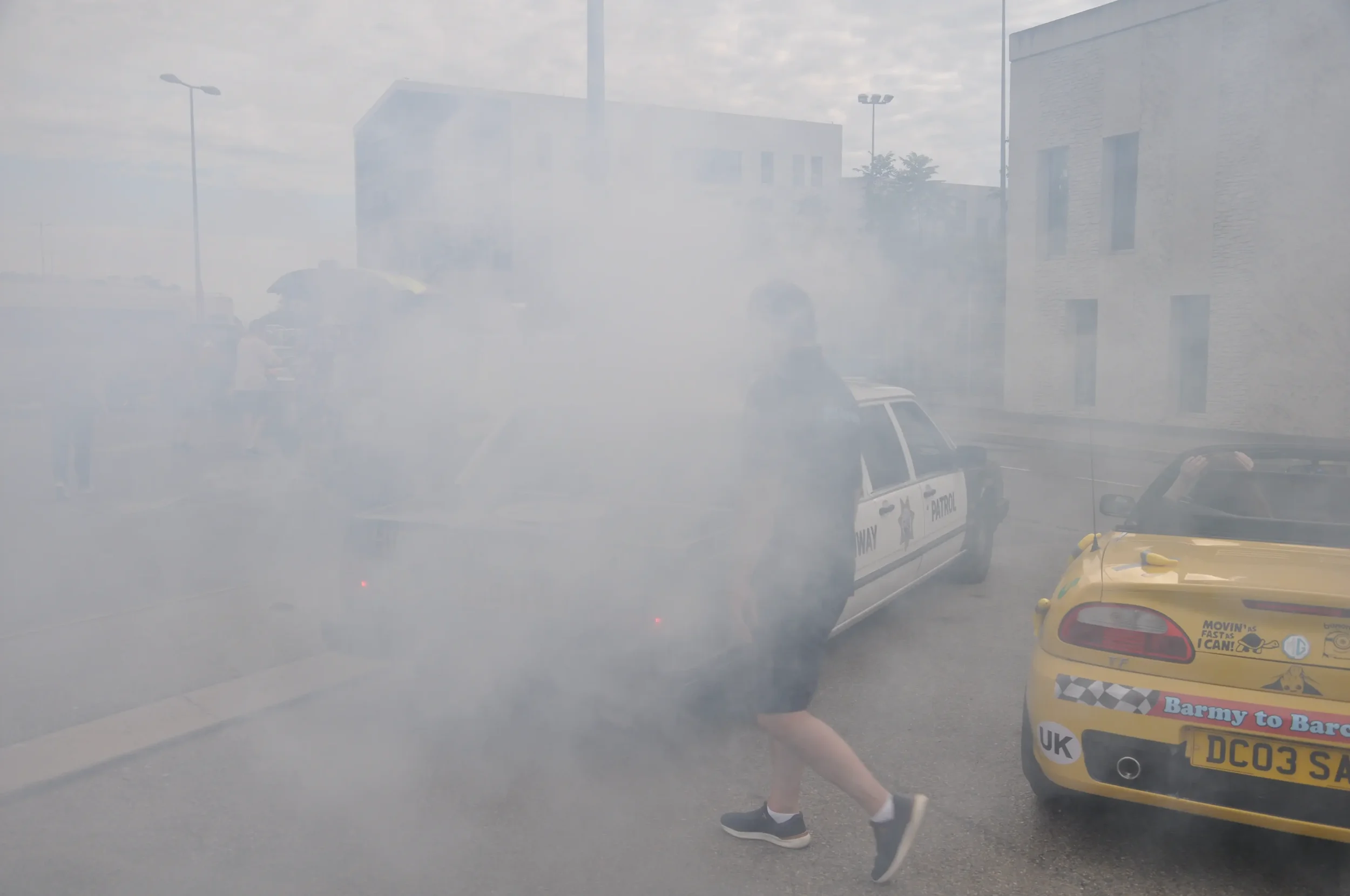 Person walking past a police car and a yellow sports car parked on a street, with smoke or fog in the air.