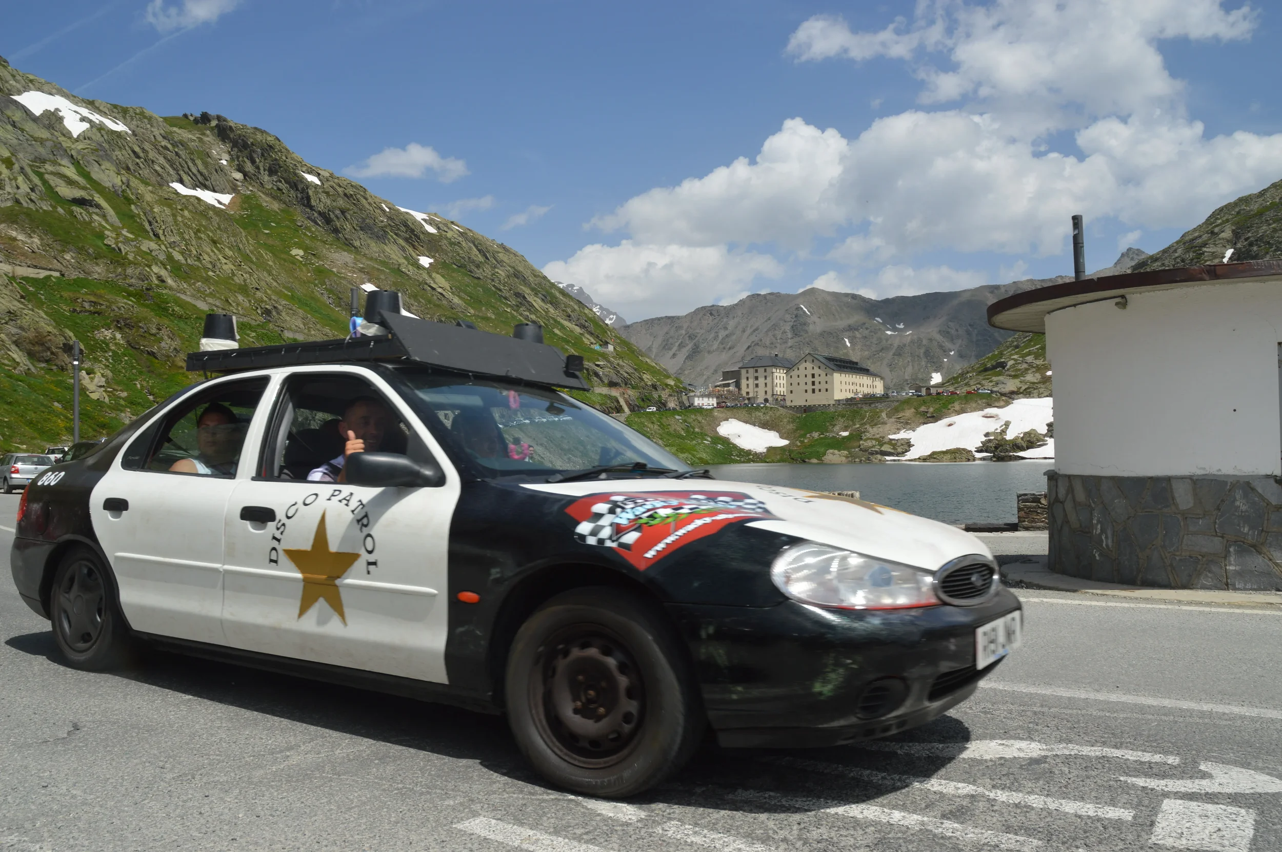 A black and white police car with a gold star emblem and the words 'DISCO PATROL' on the side, driving along a mountain road by a lake under a partly cloudy sky.