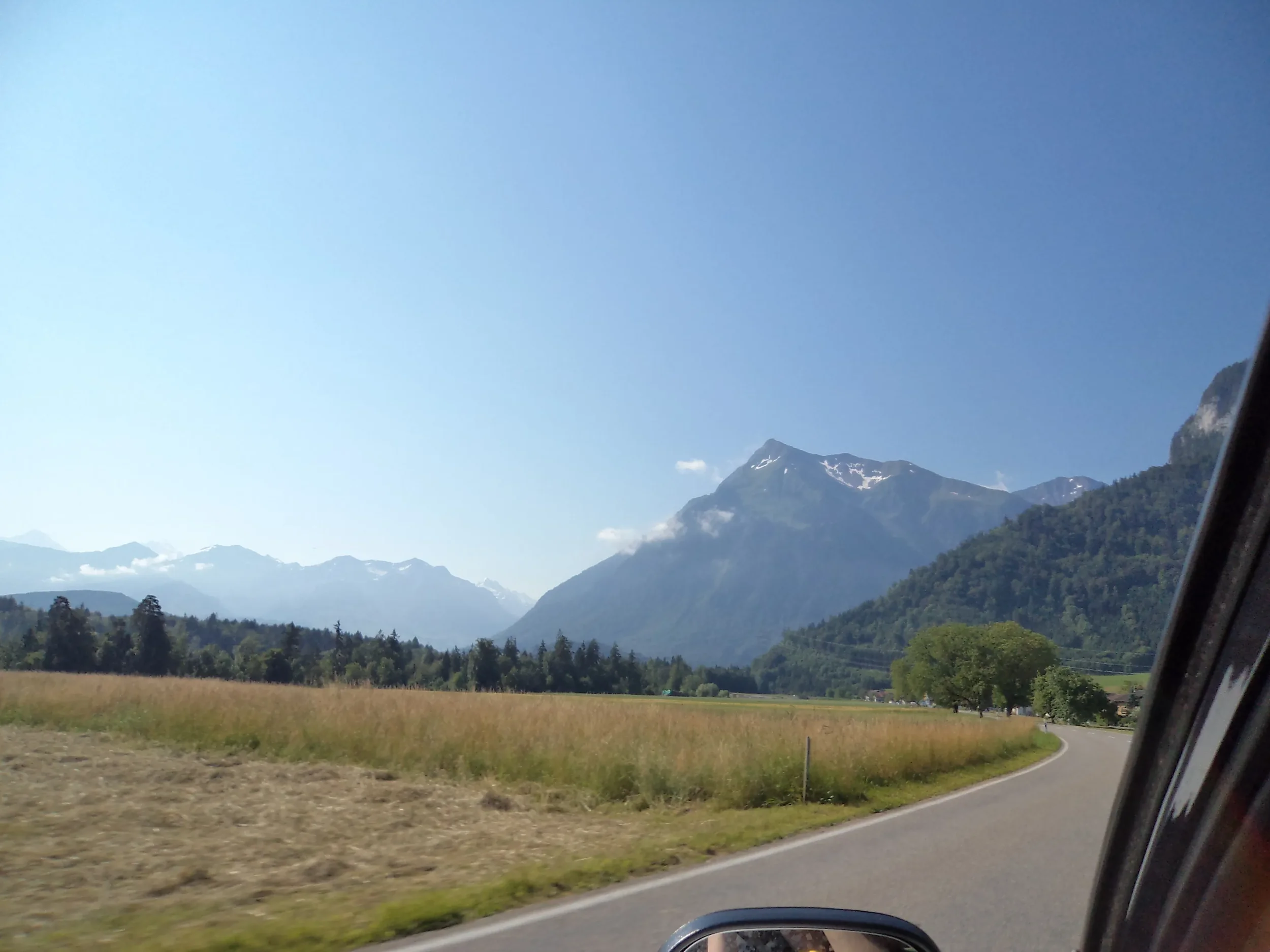 Scenic view of mountains and green fields seen from a moving car on a curved road, with part of the car's window and side mirror visible.