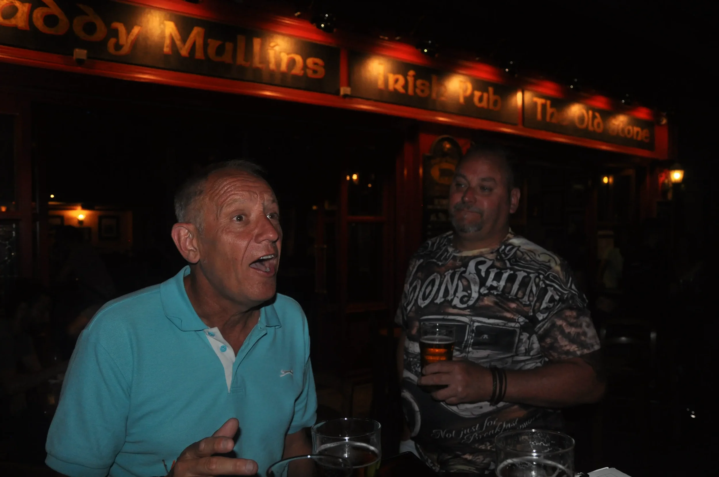 Two men talking and holding drinks in a bar. A sign with text is visible in the background.