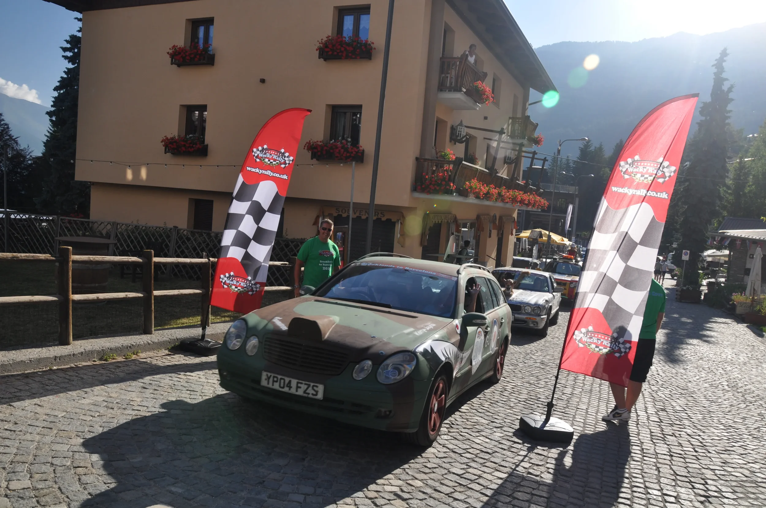 A vintage car with a camouflage paint job on a cobblestone street, flanked by two red flags with checkered patterns and Wacky Races logos, in a small town with a beige building and mountain backdrop.