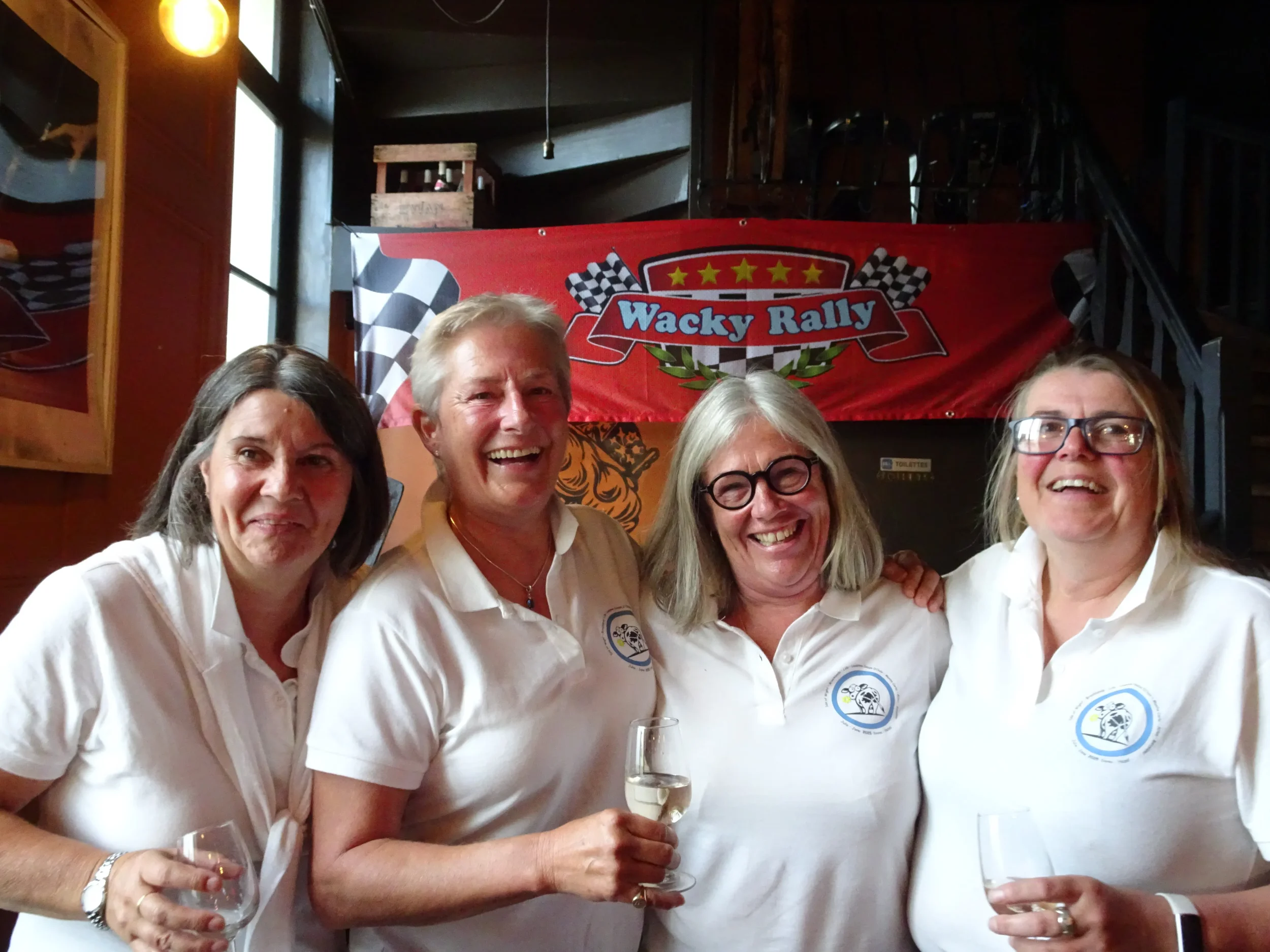Four women wearing white polo shirts smiling and holding wine glasses, standing in front of a red banner with checkered flags and the words 'Wacky Rally' inside a racing-themed design.