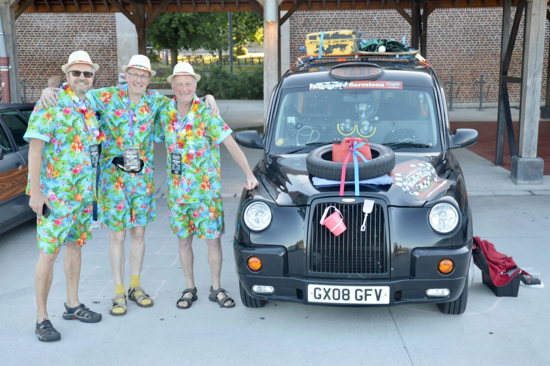 Three men dressed in colorful Hawaiian shirts, shorts, and hats standing next to a black Mini Cooper with travel and beach gear on its roof and hood, under a wooden shelter.