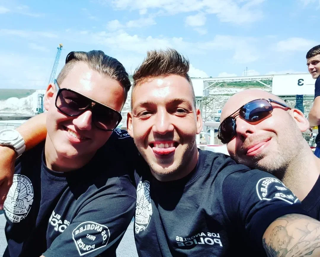 Three smiling men taking a selfie outdoors, wearing police uniforms with patches, with a roller coaster and a young man in the background.
