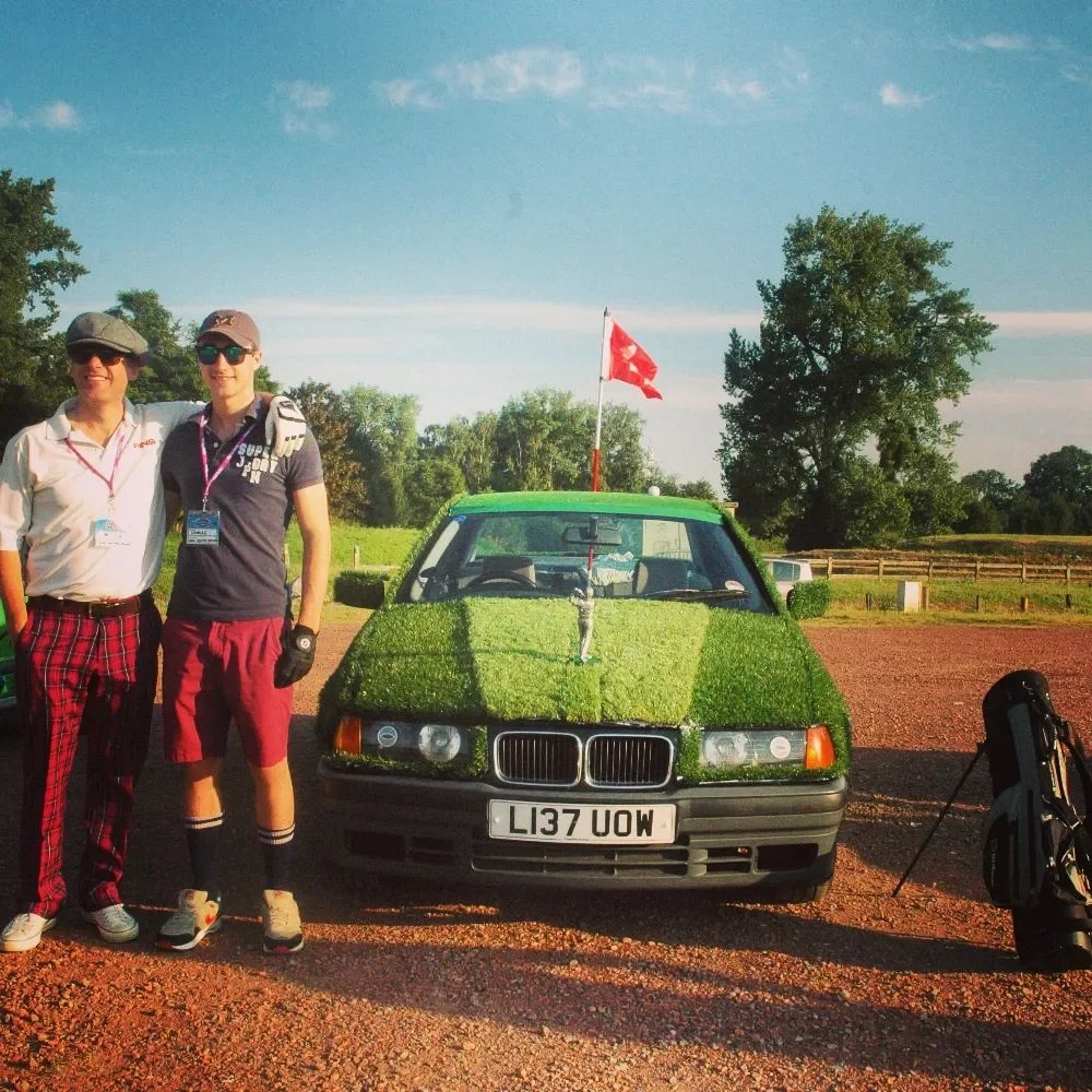 Photo of two men standing next to a modified green car with grass on its hood and a small British flag, in a rural outdoor setting.