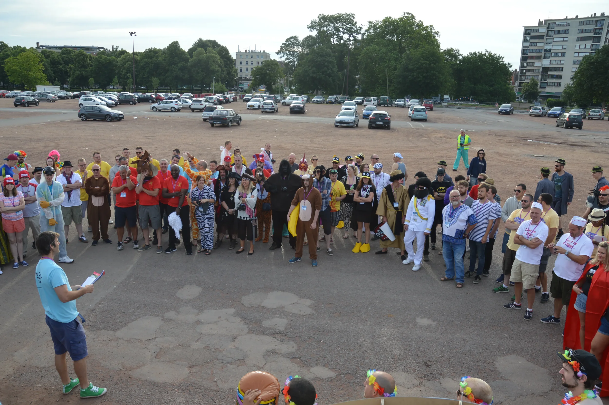 A group of people gathered outdoors, many dressed in costumes including animals, pirates, and clowns, listening to a man holding a clipboard.