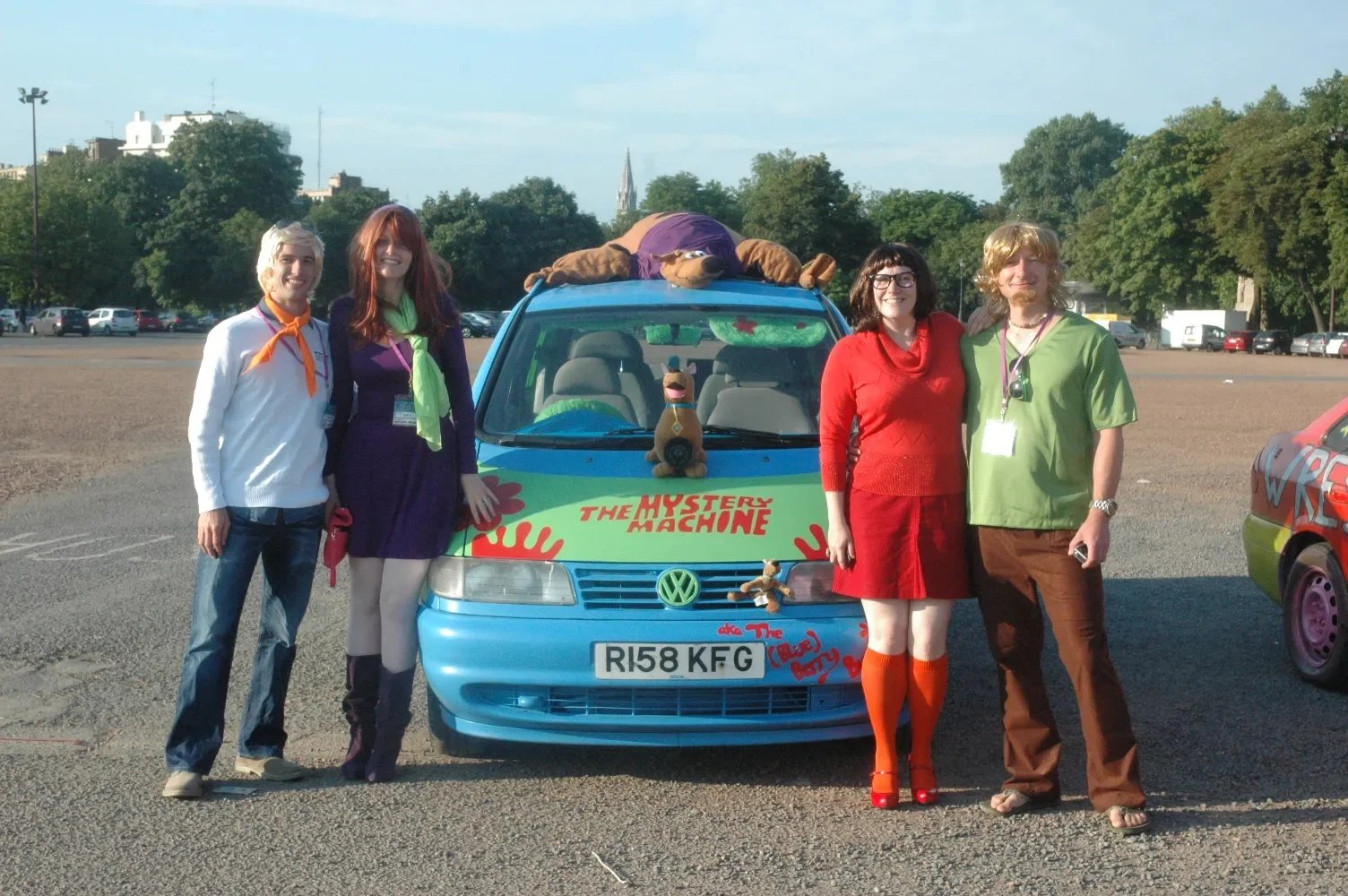 Five people dressed in colorful costumes stand in front of a blue van decorated with characters from Scooby-Doo, including Scooby-Doo plush toys on the hood and a Mystery Machine sign. The group is outdoors in a parking lot with trees and buildings i