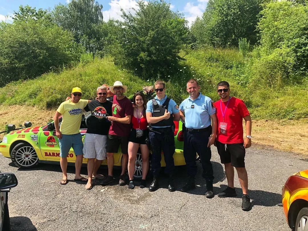 Group of seven people standing outdoors in front of a colorful Jurassic Park-themed car, smiling and posing for the photo, with green trees and clear sky in the background.