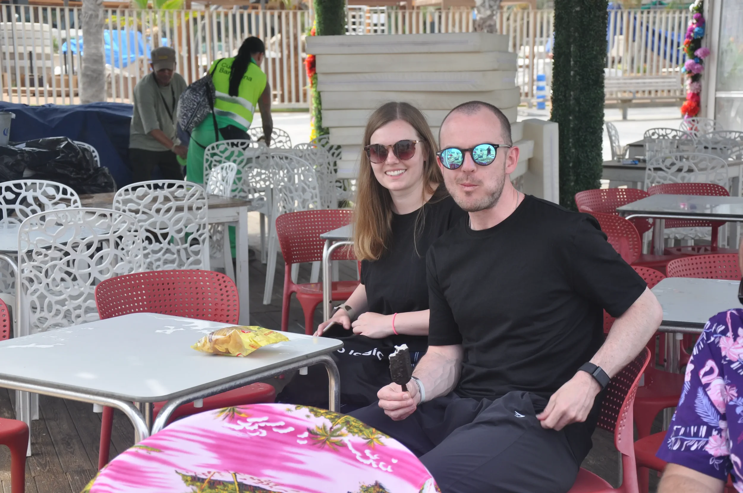 Two people sitting at a table outdoors at a restaurant, smiling at the camera, with the man holding an ice cream bar. The woman has long brown hair and is wearing sunglasses and a black shirt. The man has a shaved head, sunglasses, and a black shirt.