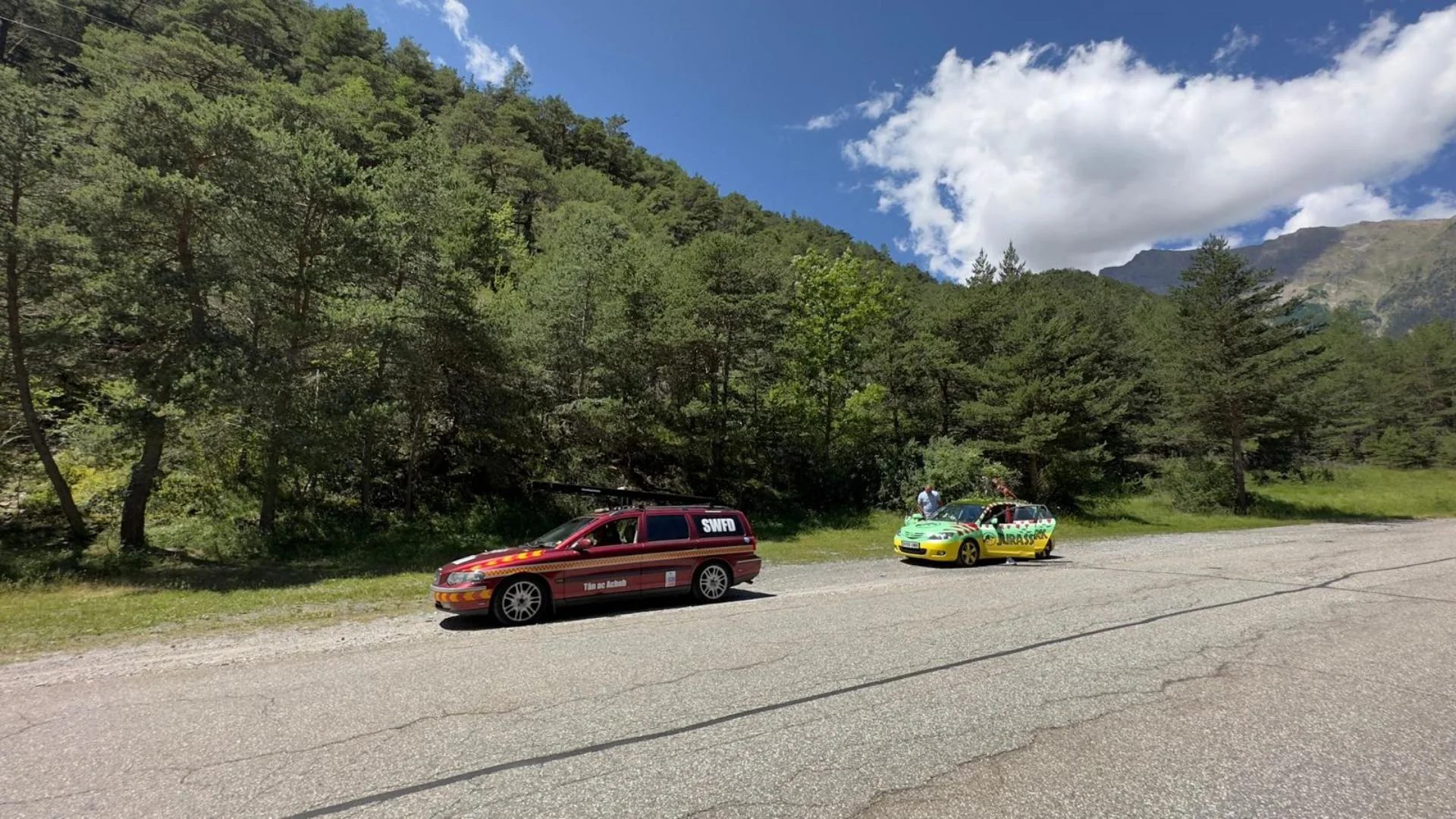 Two small cars, one red and one yellow-green, parked on the side of a mountain road with trees and mountains in the background.