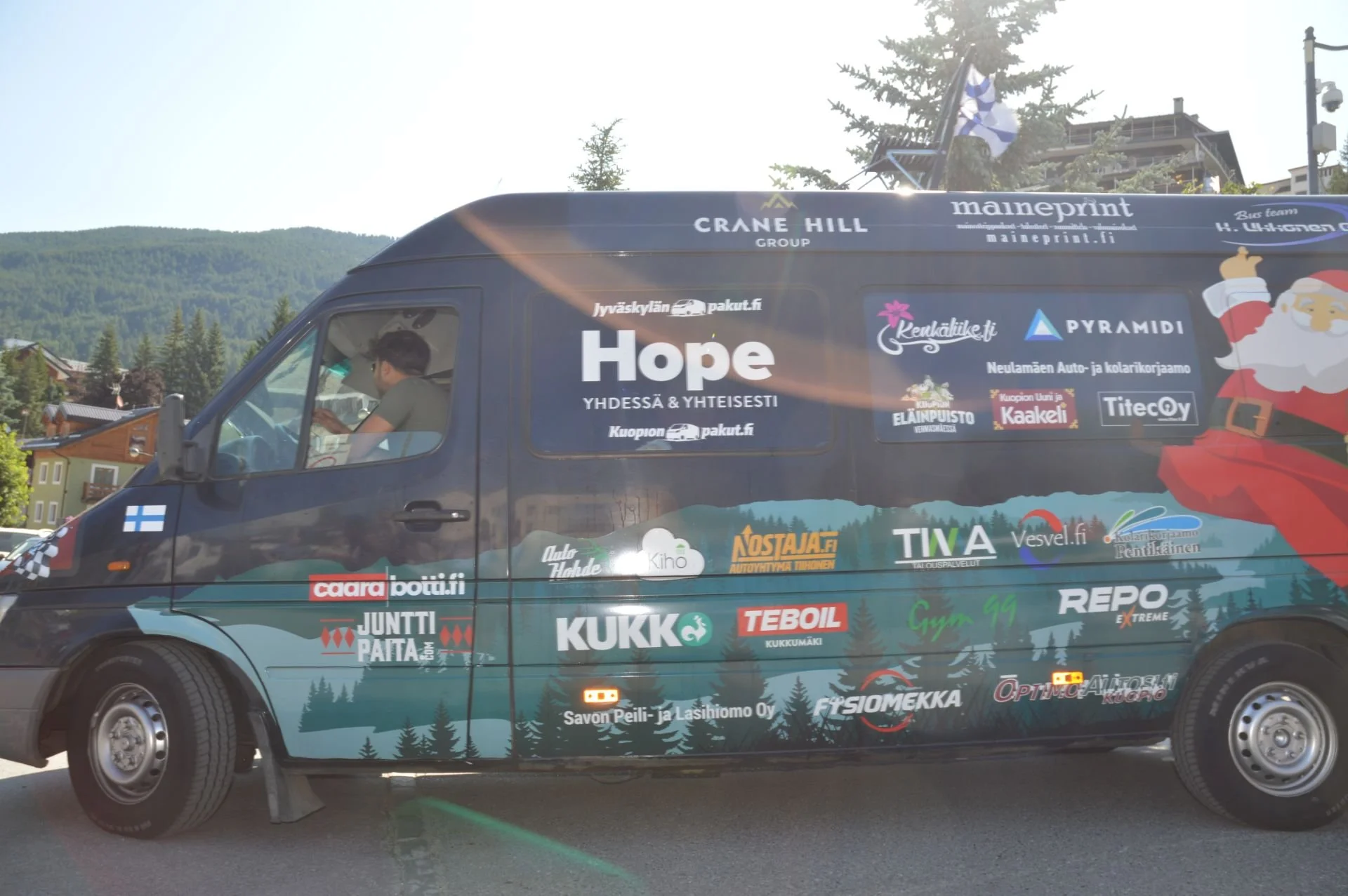 Black van decorated with various company logos and a Santa Claus illustration, parked outdoors with mountains and trees in the background, and a Finnish flag sticker on the door.