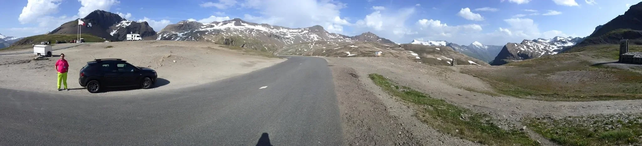 A mountain landscape with snow-capped peaks, a paved road, a person in bright clothing standing next to a black car, and a few scattered vehicles and structures.