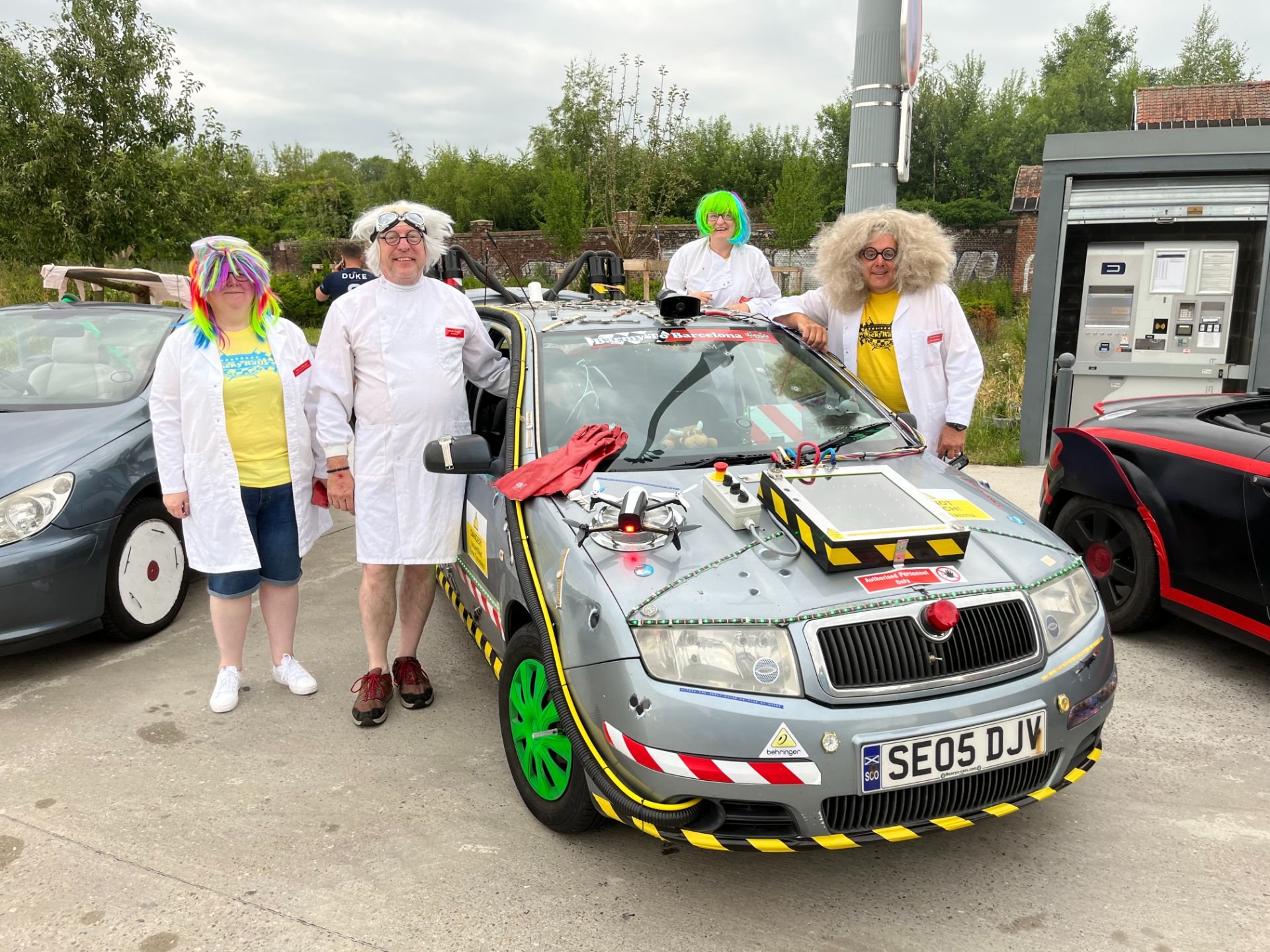 Four people dressed as scientists with colorful wigs and white lab coats around a small car fitted with various electronic equipment and safety markings, indicating a science or engineering event outdoors.