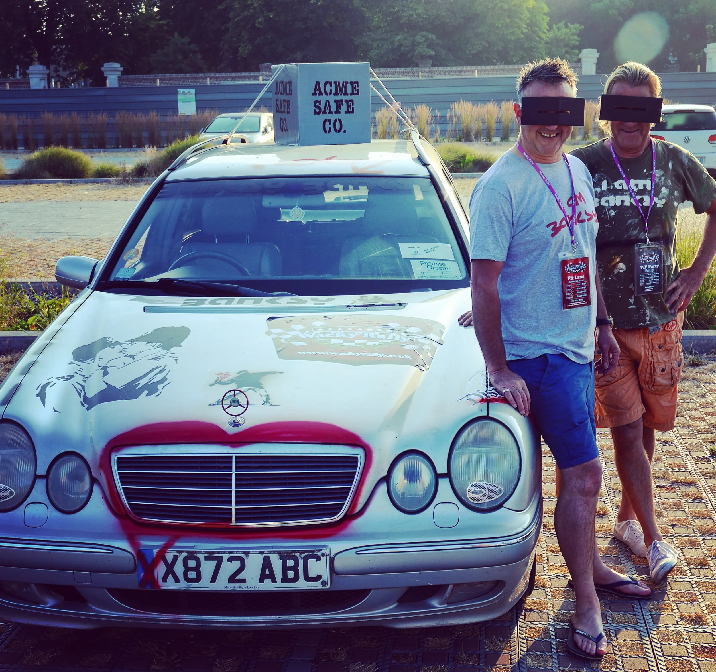 A decorated silver Mercedes-Benz car with graffiti-style art on the hood, parked outdoors with two smiling men standing beside it, wearing glasses covered with black tape, and badges around their necks.