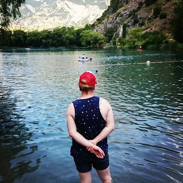 Person standing at the edge of a river with their back to the camera, wearing a red cap, a sleeveless navy top with polka dots, and black shorts, surrounded by trees and mountains.
