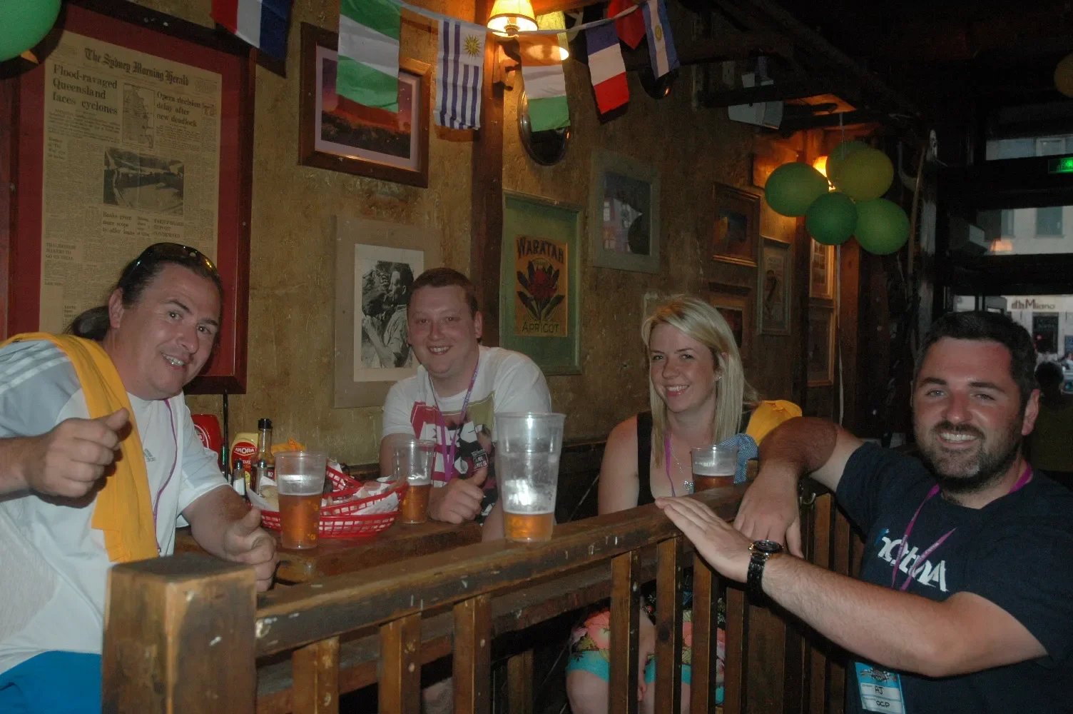Four friends sitting at a bar with drinks, smiling, in a decorated pub with flags and pictures on the wall.