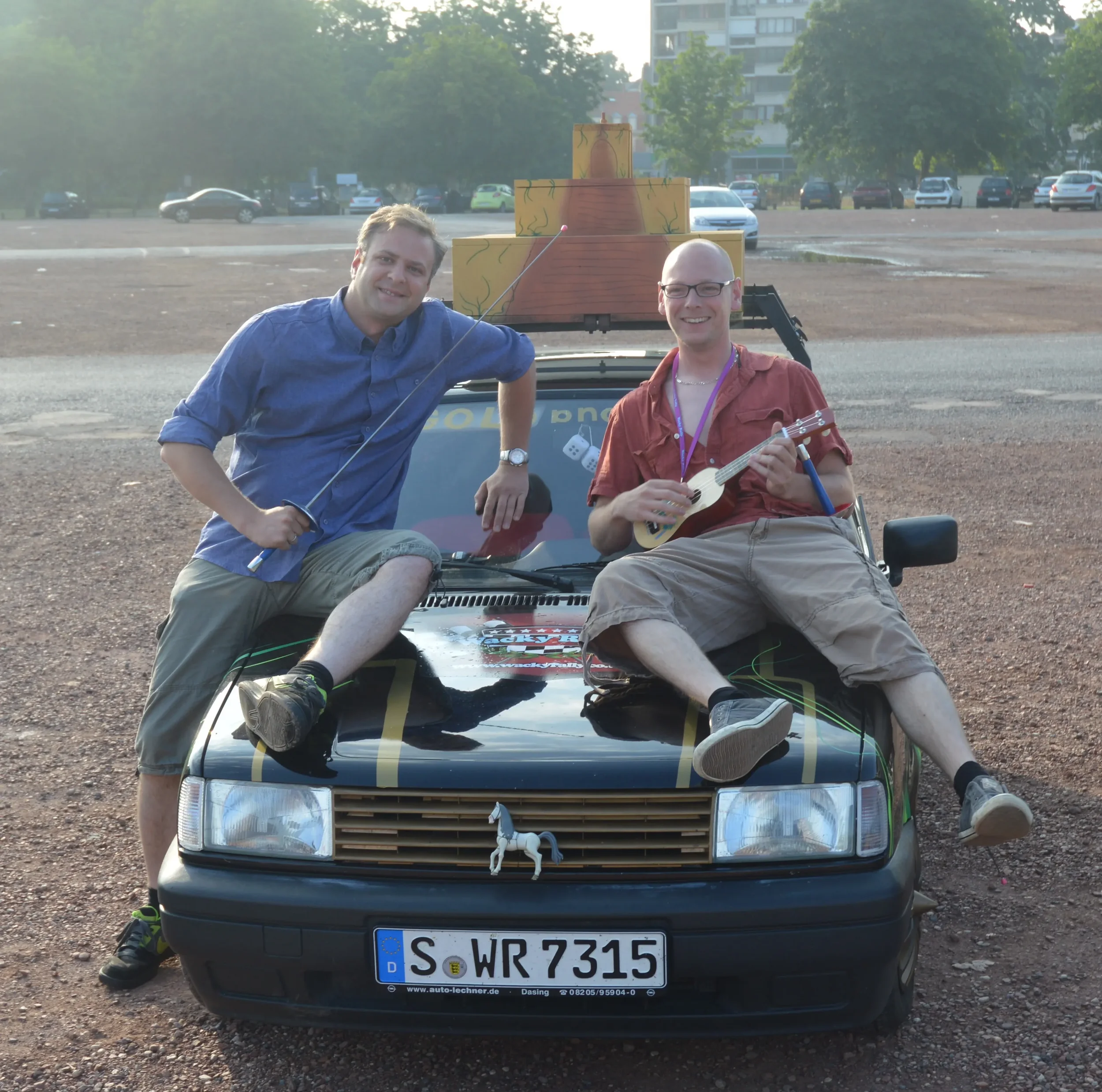 Two men with short hair sitting on the hood of a black car, both smiling, one holding a sword and the other holding a ukulele, in an open parking lot.