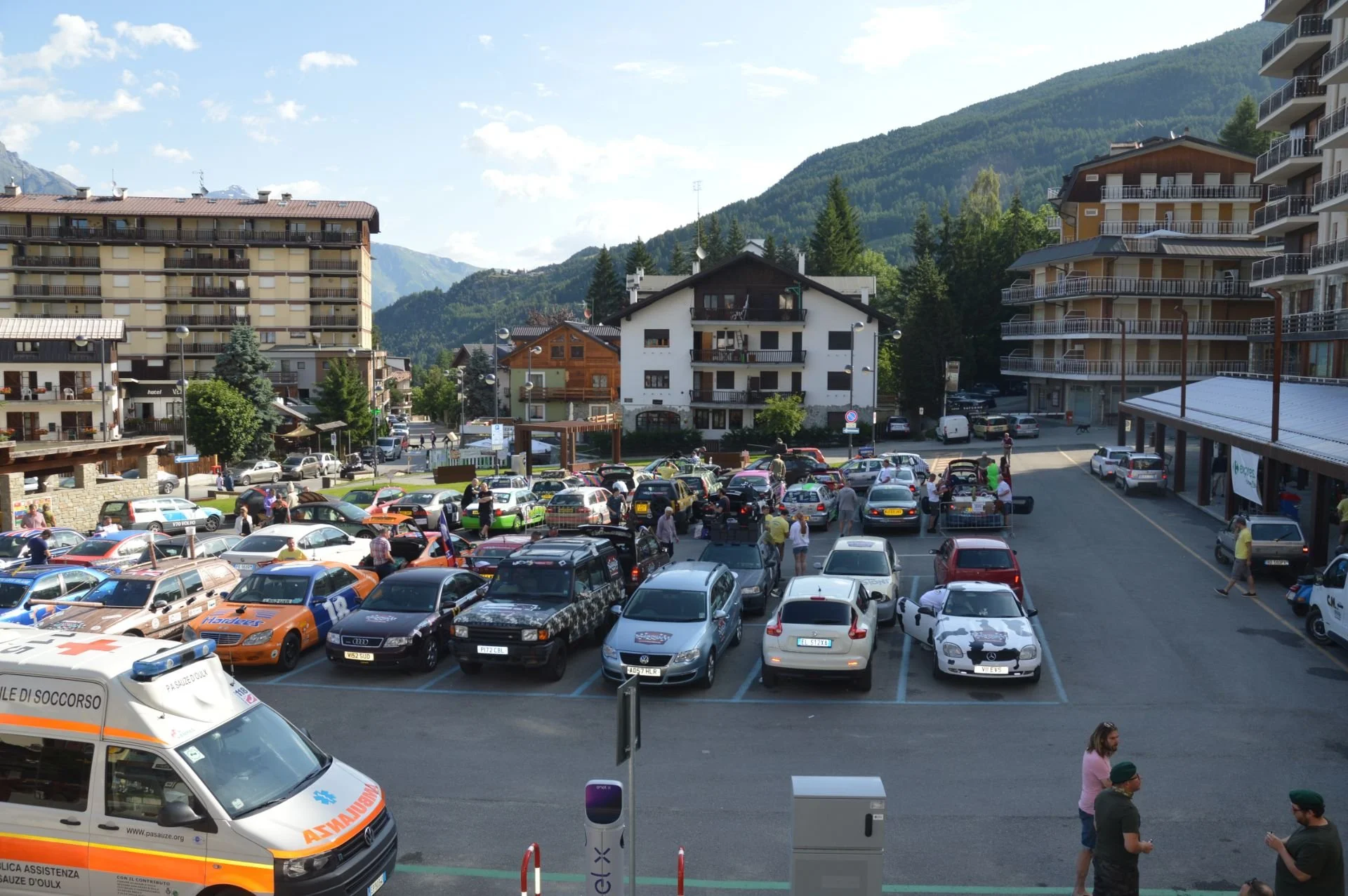 A parking lot filled with various cars, some with open trunks, in front of buildings and mountains in the background on a sunny day. Several people are walking or standing near the cars, and an ambulance is visible in the foreground.