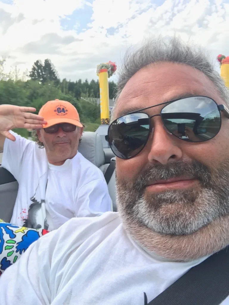 Two men are taking a selfie outside in a convertible car during the daytime. One man has gray hair, a beard, and is wearing sunglasses and a white shirt. The other man is wearing a bright orange cap, sunglasses, and a white T-shirt with a graphic. Th