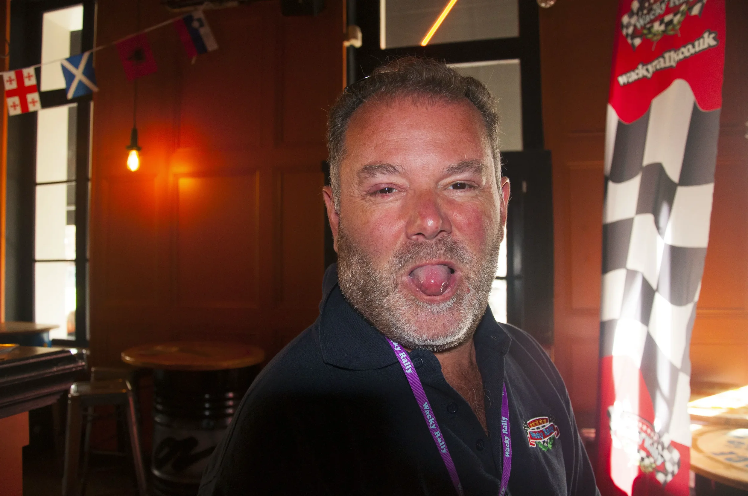 A man with short gray hair and a beard, making a playful face with his tongue out, in a room decorated with flags and racing-themed decorations.