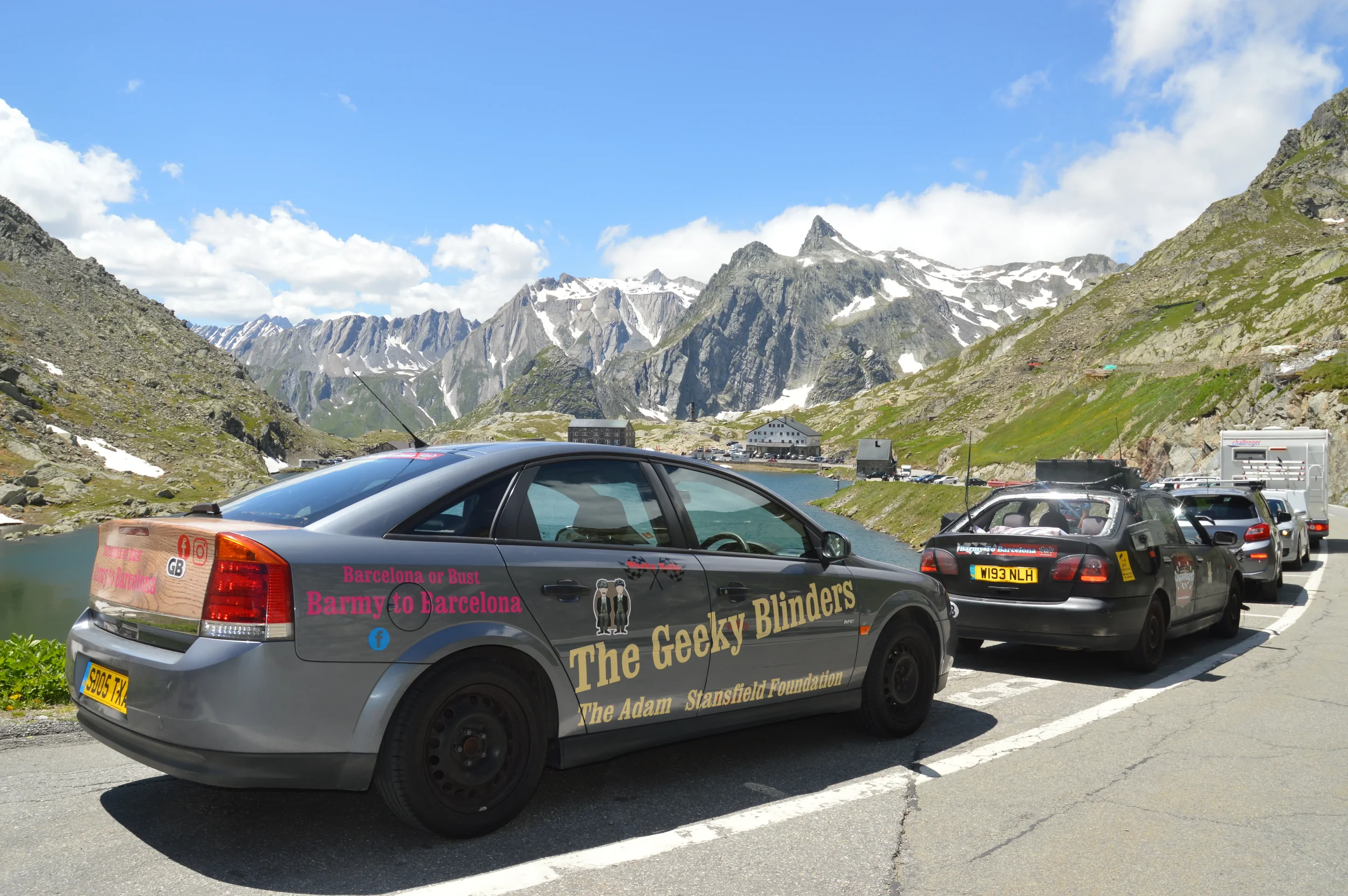 Line of cars parked along a mountain road near a lake with snow-capped peaks in the background. The closest car is a gray vehicle with the text 'The Geeky Blinders' and other references to Barcelona and Barmy.