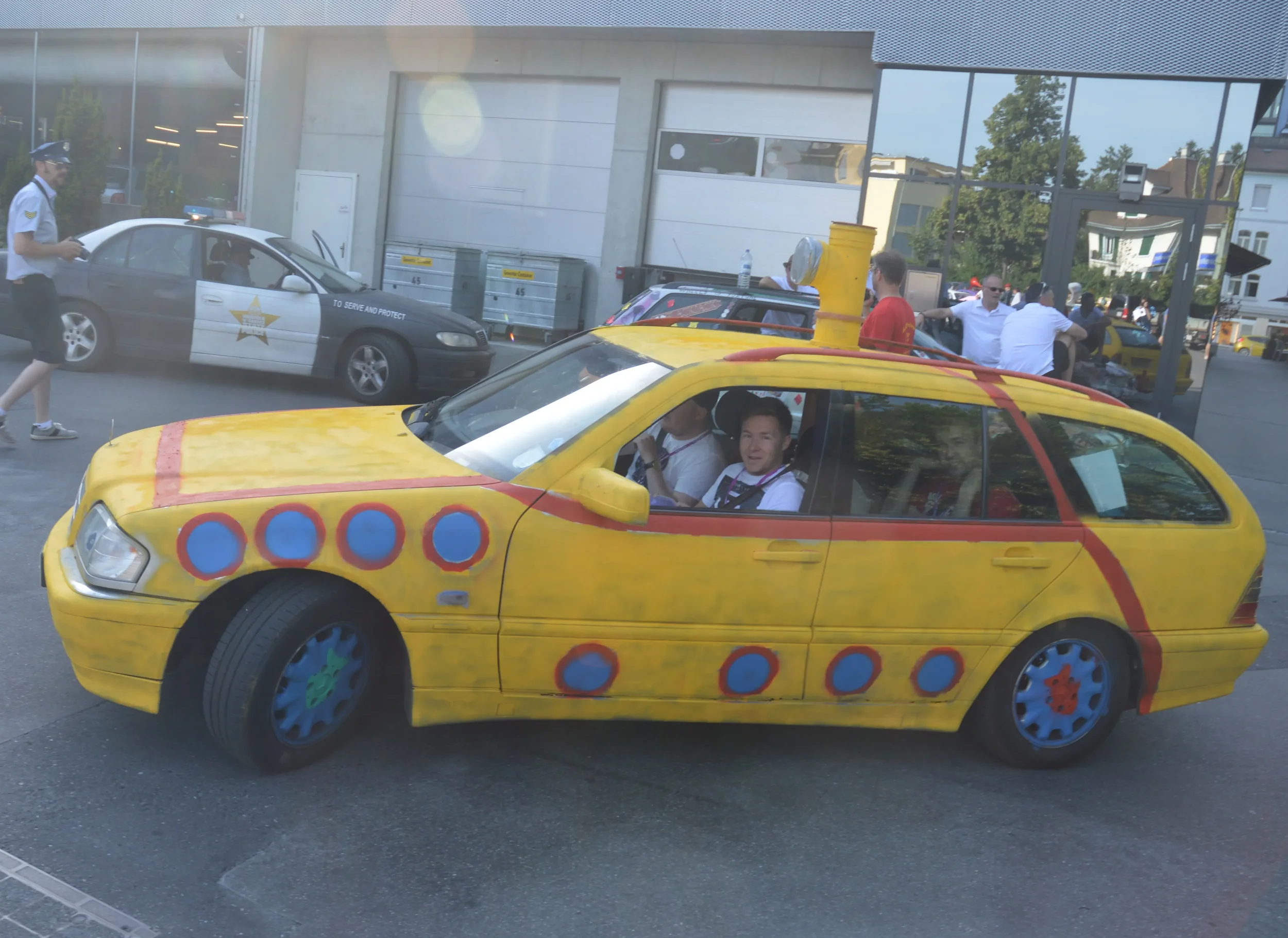 A yellow car with colorful, whimsical paint resembling a child's drawing, parked outside a building. People are gathered around, some seated inside the car, which has painted wheels and a chimney on the roof.