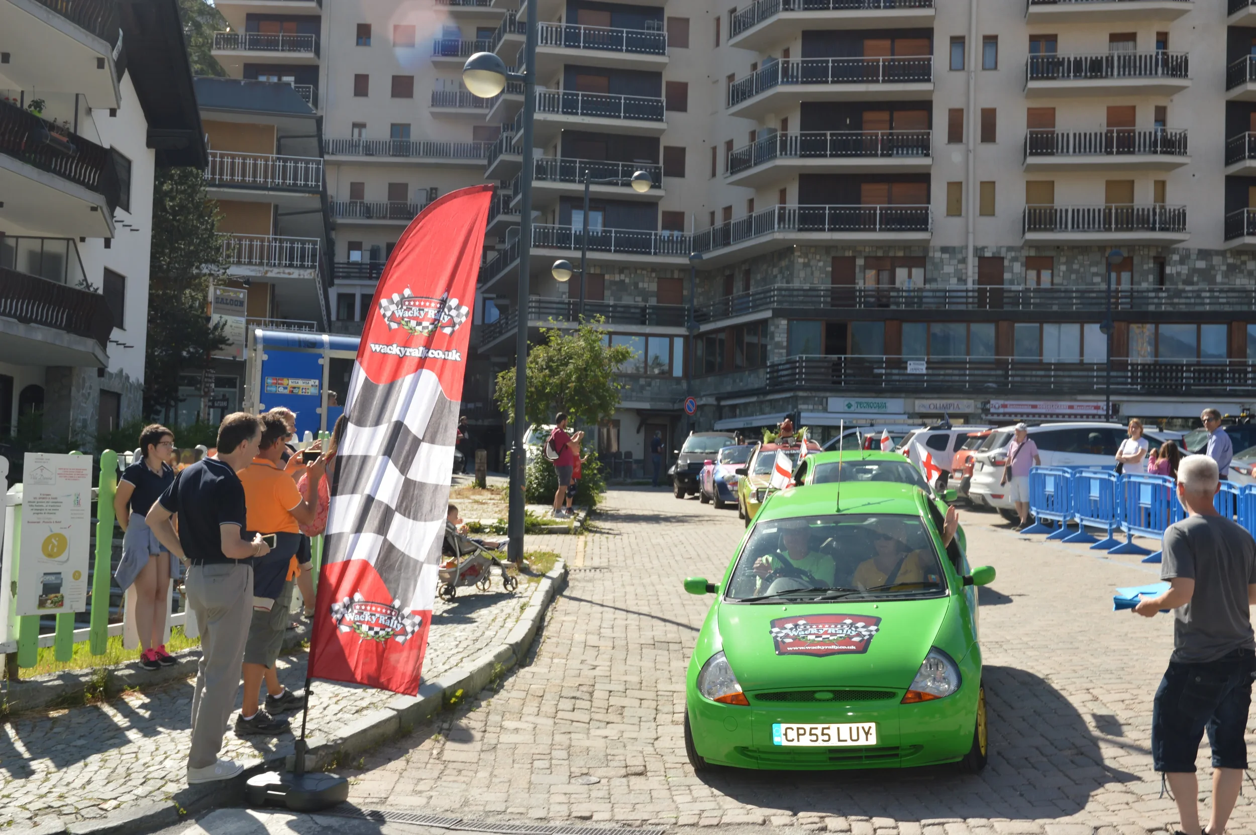 A green car with a race-related sticker on the hood parked on a cobblestone street during a car rally event. A red checkered flag banner with a racing logo is beside the car. People are gathered around, some taking photos and others observing the sce