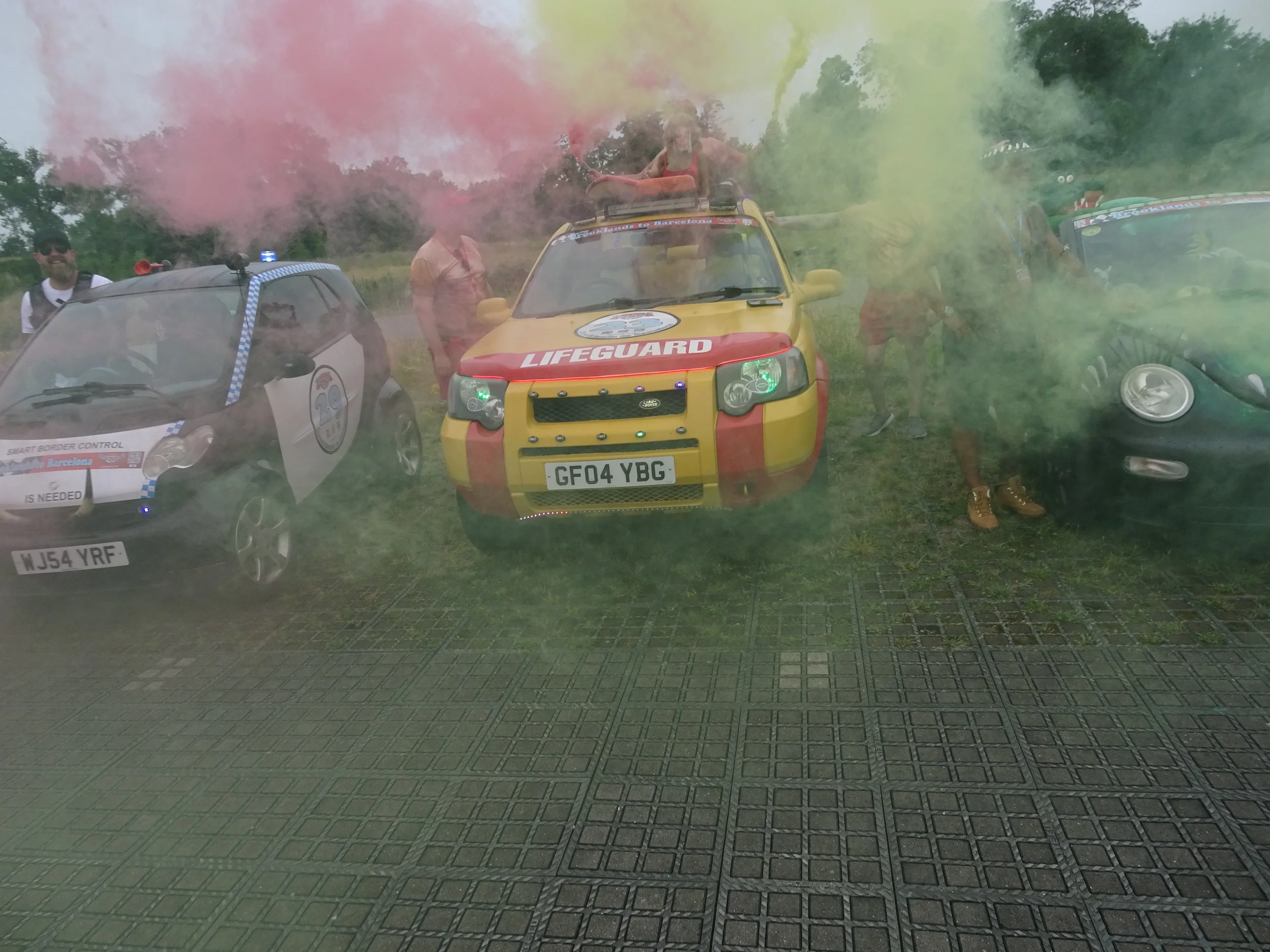 People standing behind three cars, with colored smoke in the air, outdoors on grassy ground.