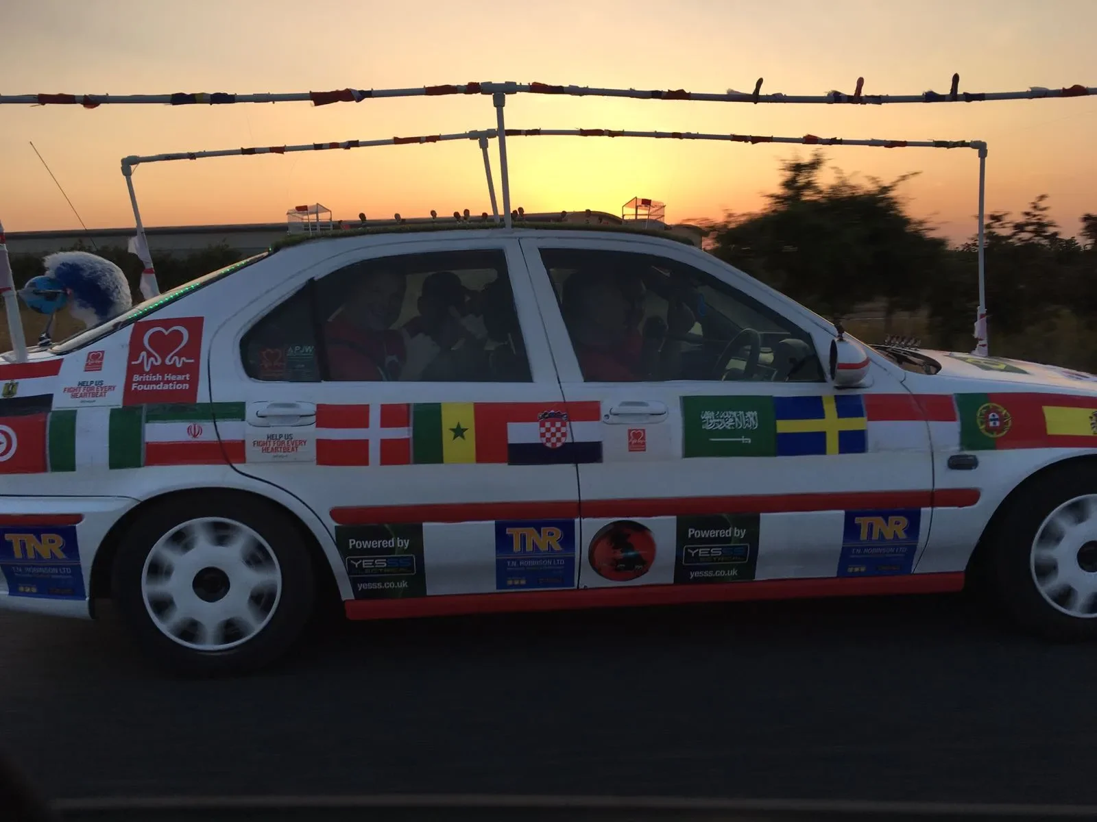 A race car decorated with international flags and sponsorship logos drives through a sunset landscape, with a person in a racing suit inside.