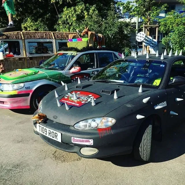Two decorated cars with various colorful decorations and props parked outdoors, with trees and a fence in the background.