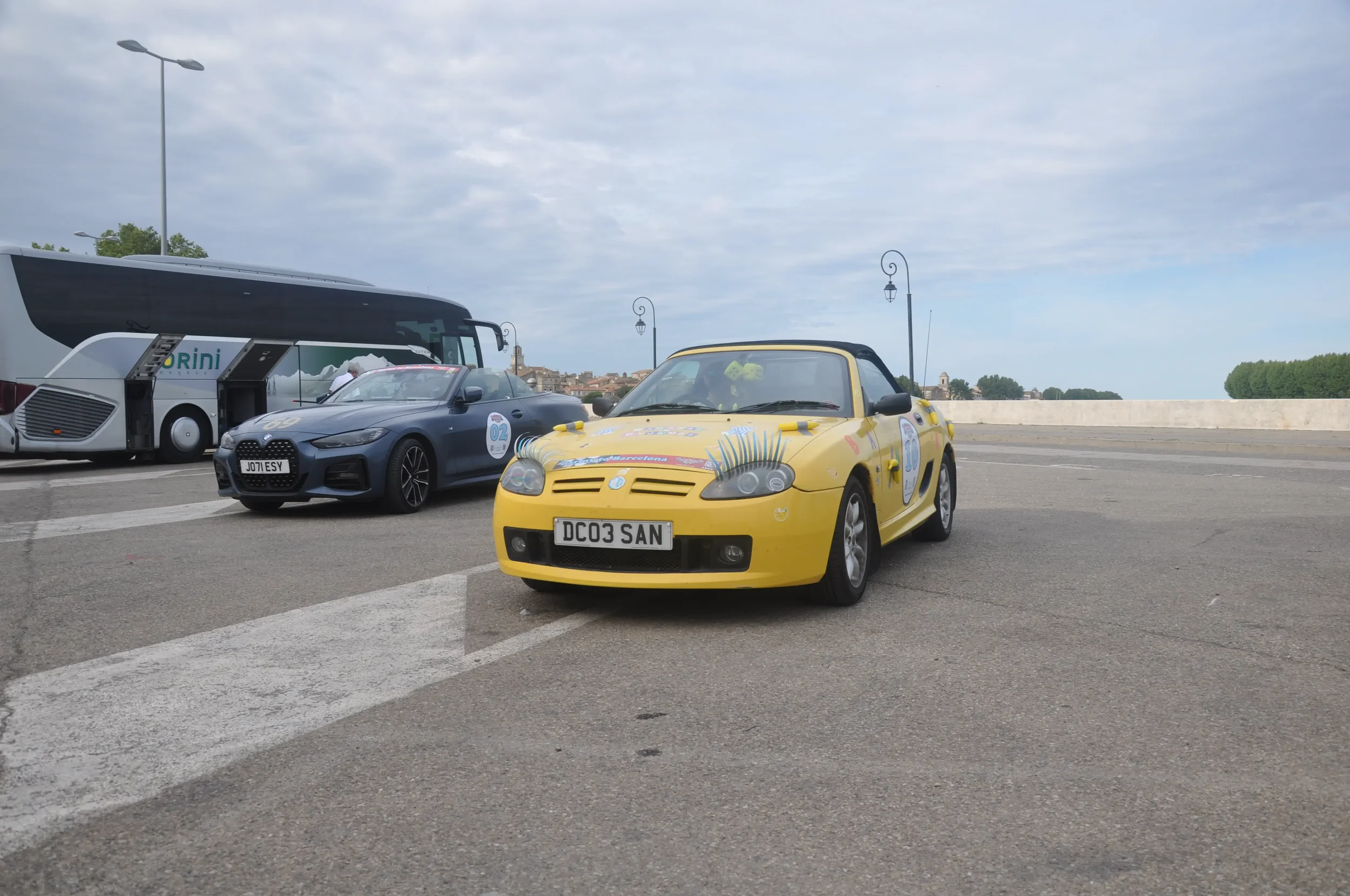 Three cars parked on a paved area with street lamps and cloudy sky, featuring a yellow convertible, a gray sports car, and a white bus in the background.