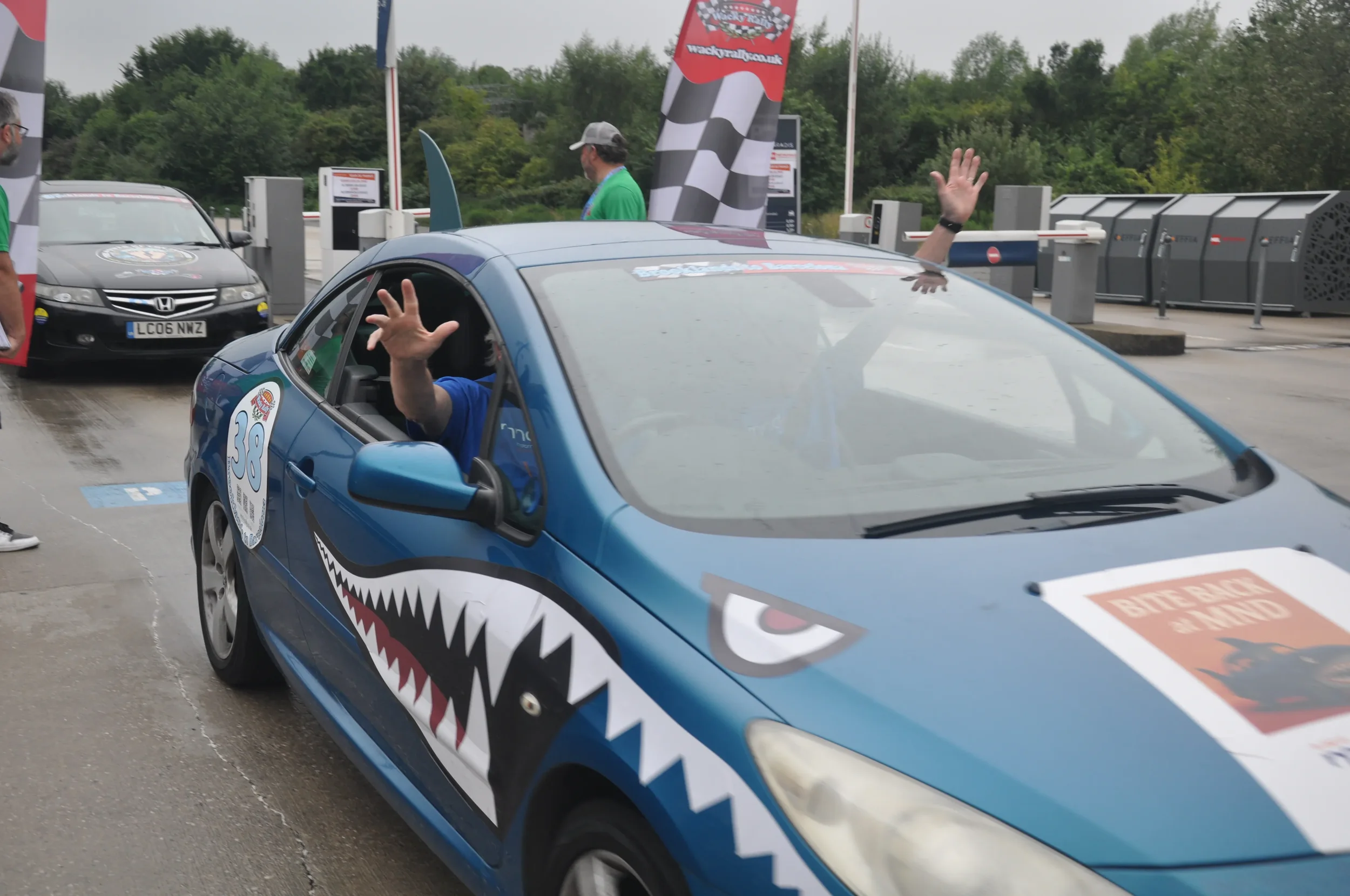 A blue race car with a shark mouth design on the side, number 38 on the door, and a 'BITE BACK & MIND' sign on the windshield, parked at a rest stop with people around it. The driver is waving through the window.