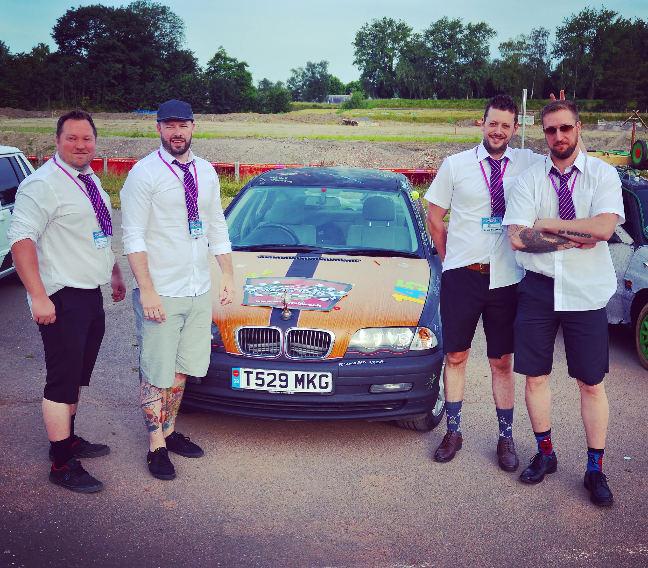Five men dressed in white shirts, black shorts, and striped ties posing in front of a decorated car with a custom license plate, at what appears to be a racing event, with an open field and trees in the background.