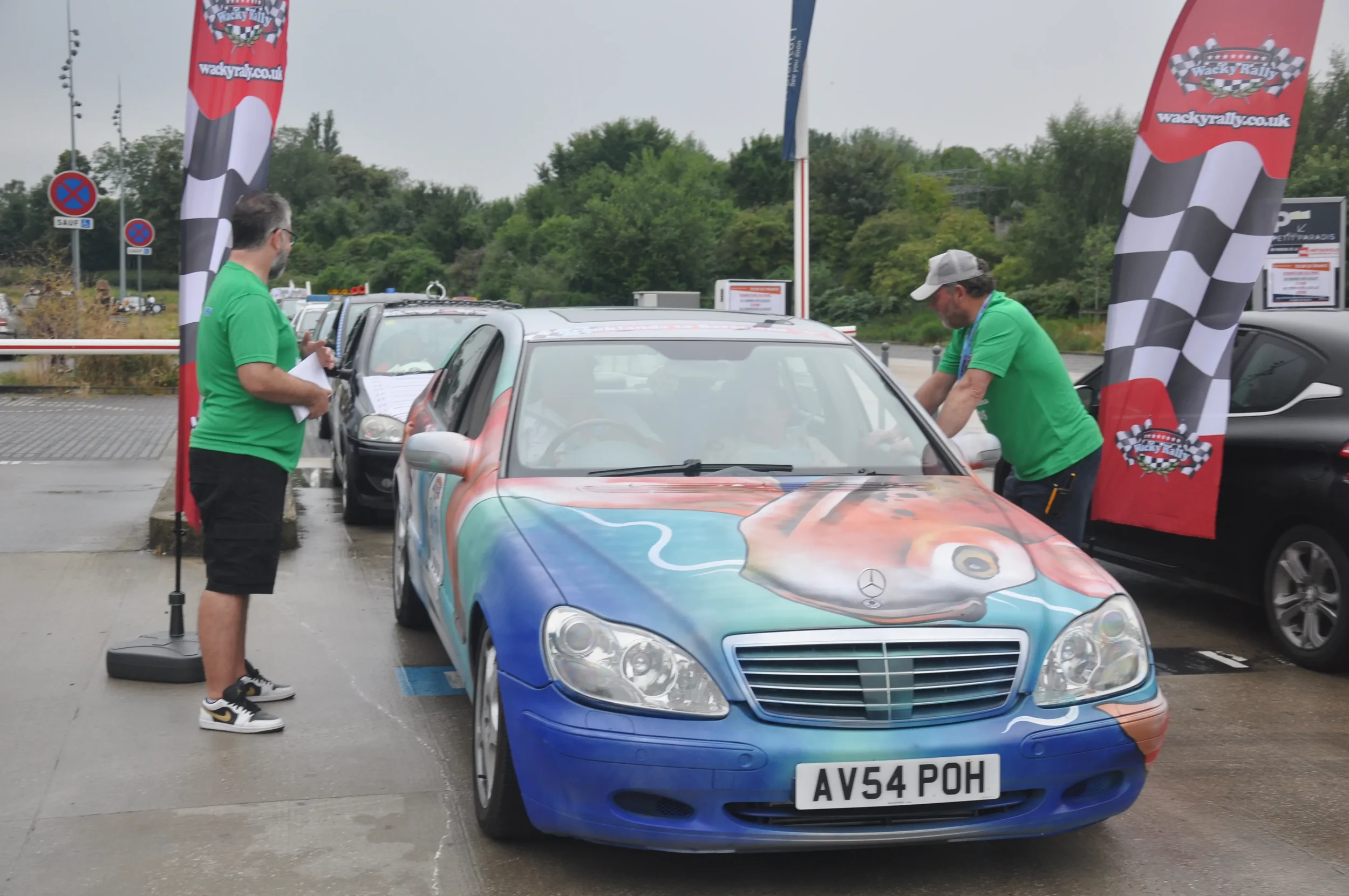 A colorful Mercedes-Benz with a racing-themed wrap being checked in a parking lot at a rally event, with flags and other cars nearby, and two men in green shirts inspecting the car.